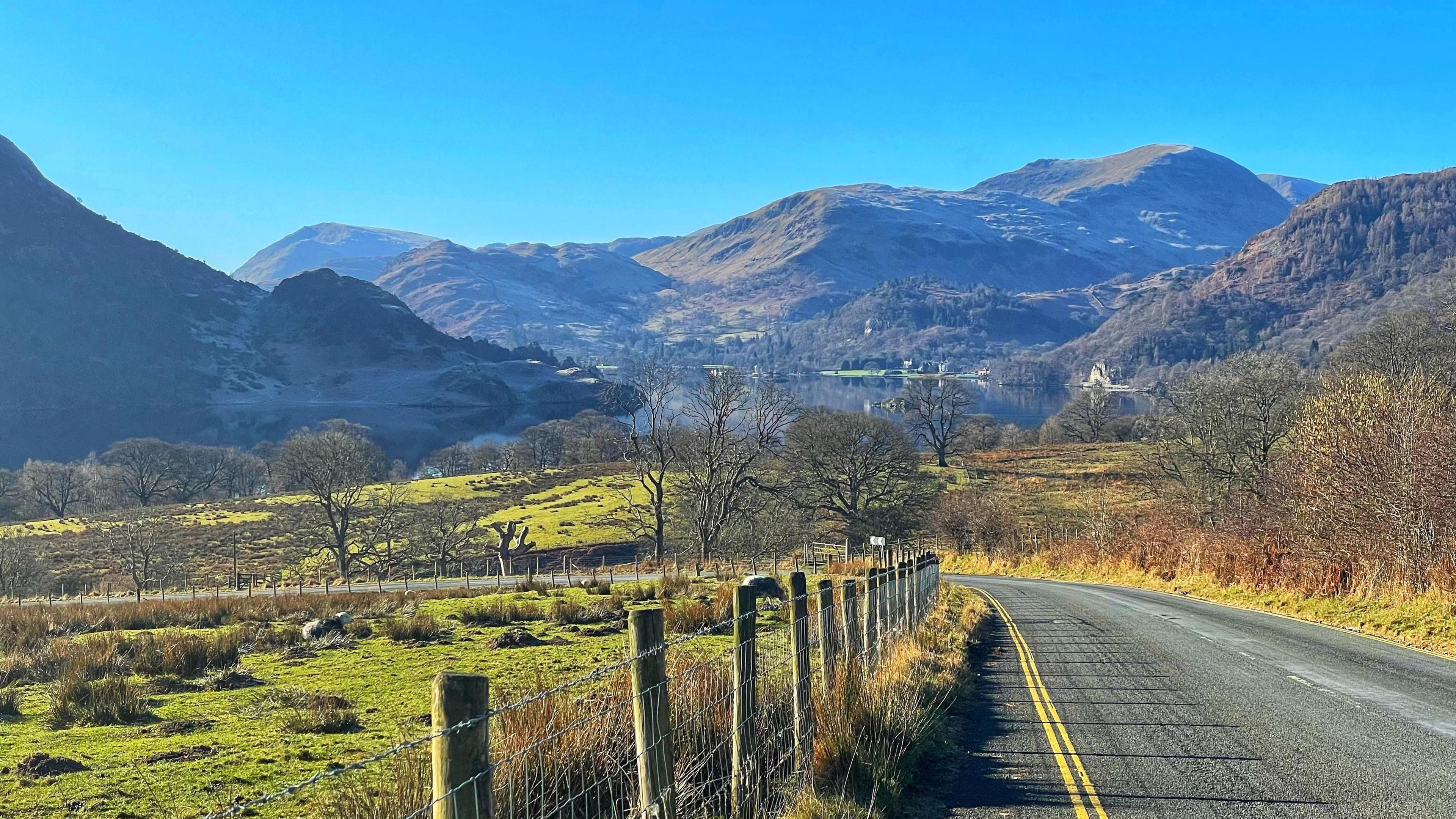 A road leading to Ullswater lake on a sunny day, with fields in the foreground and mountains surrounding the lake