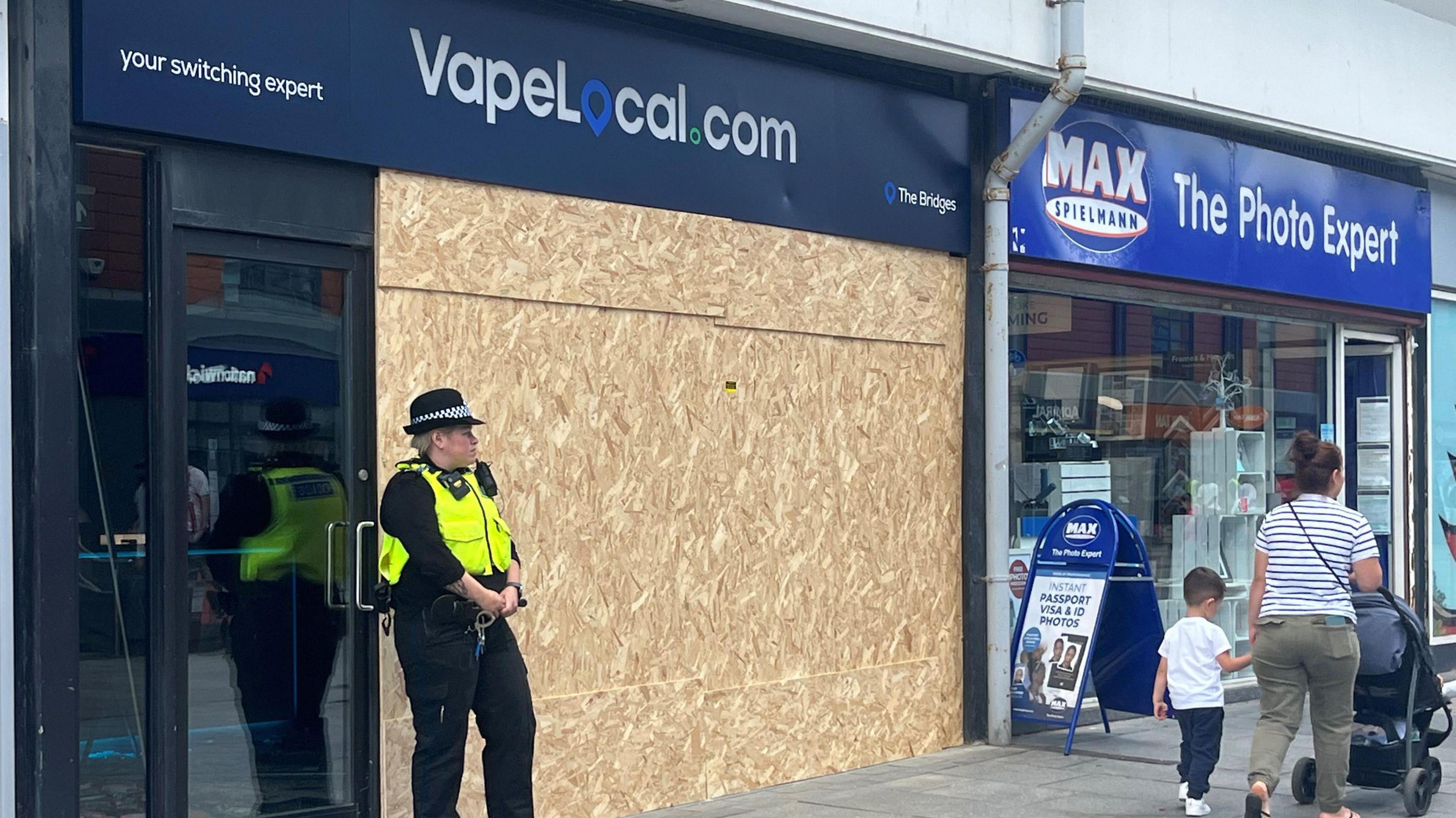 A female police officer in a black uniform wearing a black hat and yellow/green high visibility jacket stands outside a vape shop which has a large wooden board across its front.