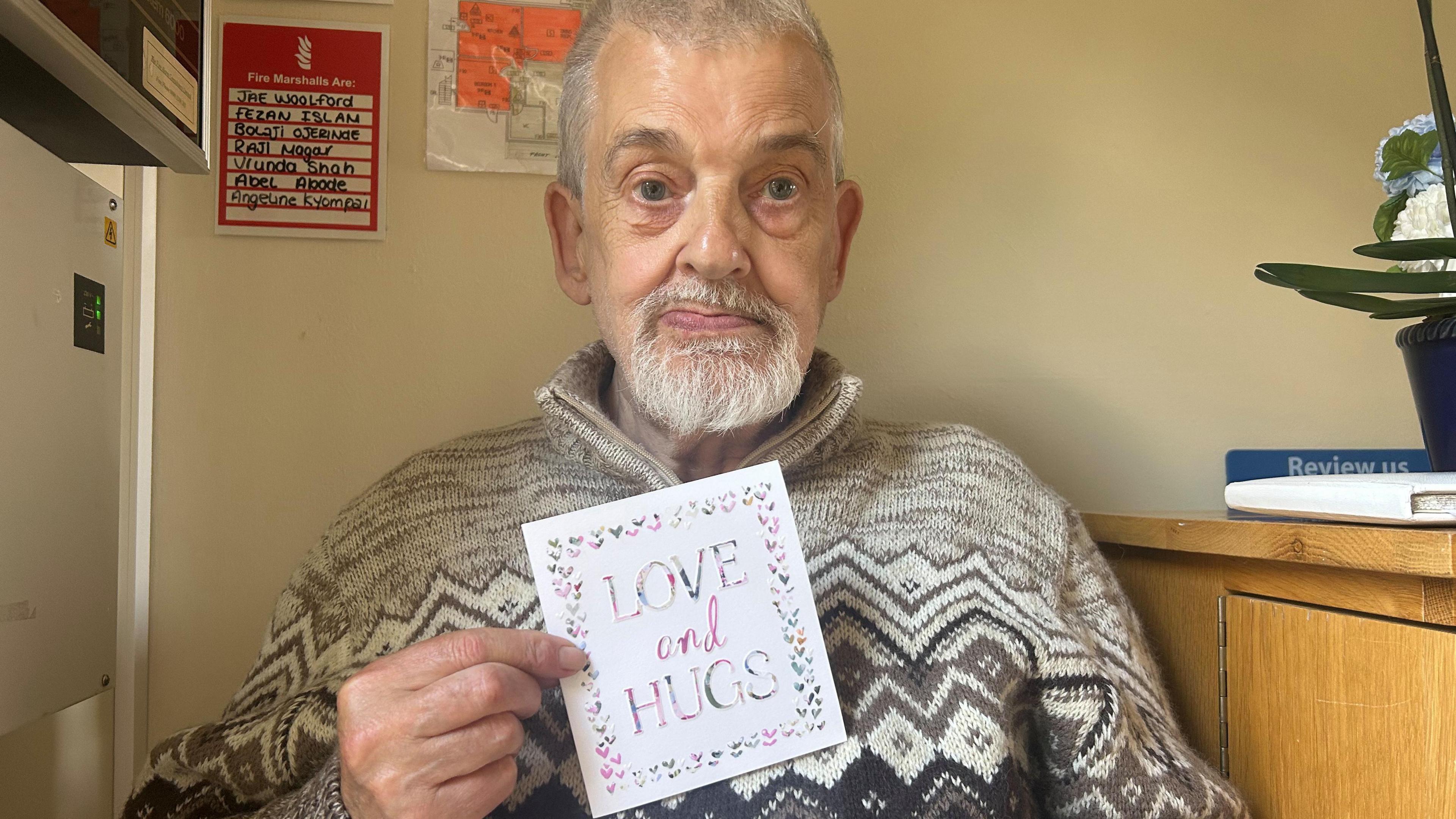 Roger from Grove Hill Care Home sits in a chair holding his card. He has short grey hair and a beard. His card says 'love and hugs', with a heart border surrounding it. Next to him is a wooden cabinet with a white book on top and a pot of white and blue flowers.