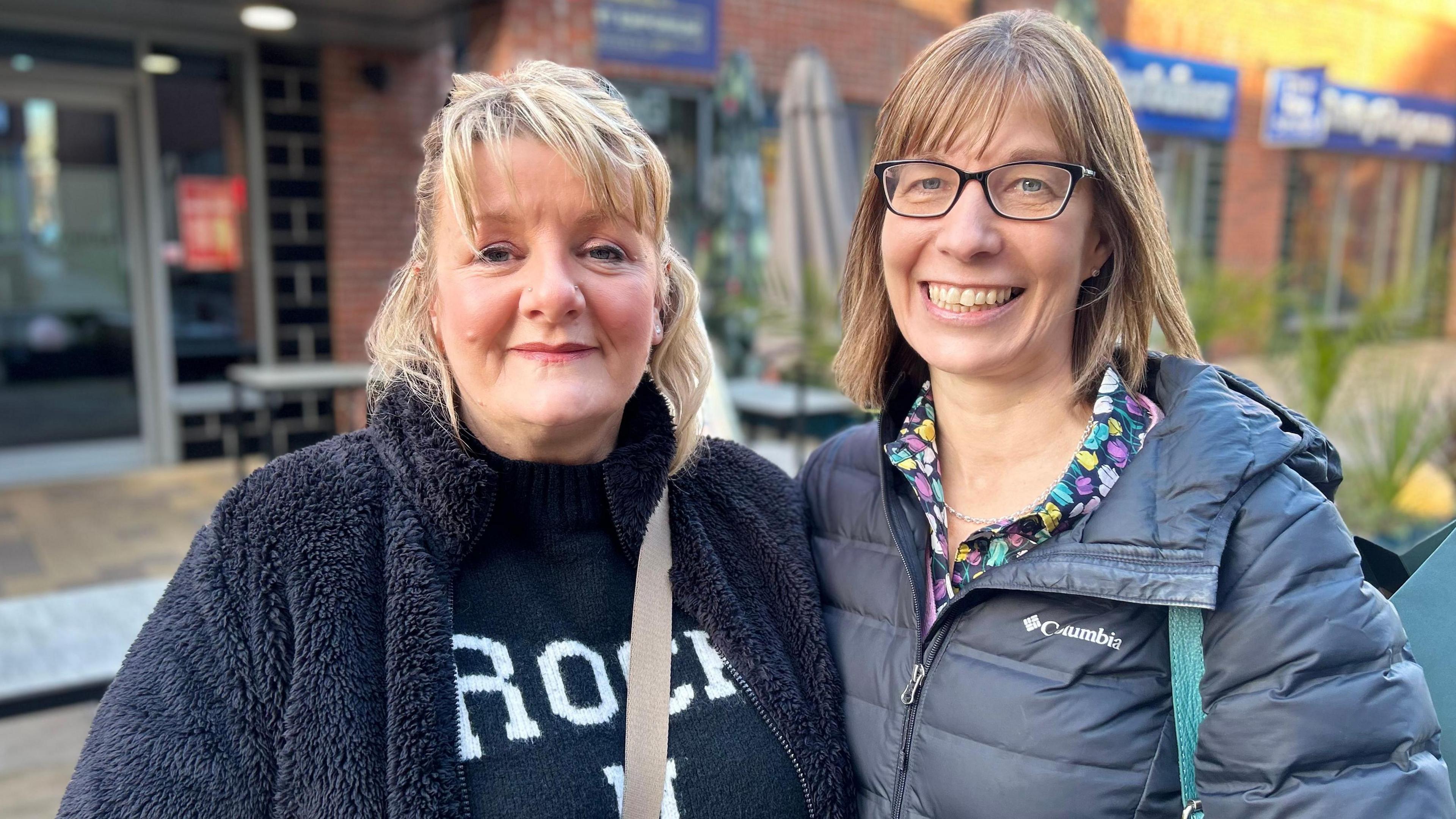 Two women standing in a street smiling into a camera. On the left, a woman with short blonde hair and a black coat is stood next to a woman with short light brown hair with and black-rimmed glasses and a blue coat.