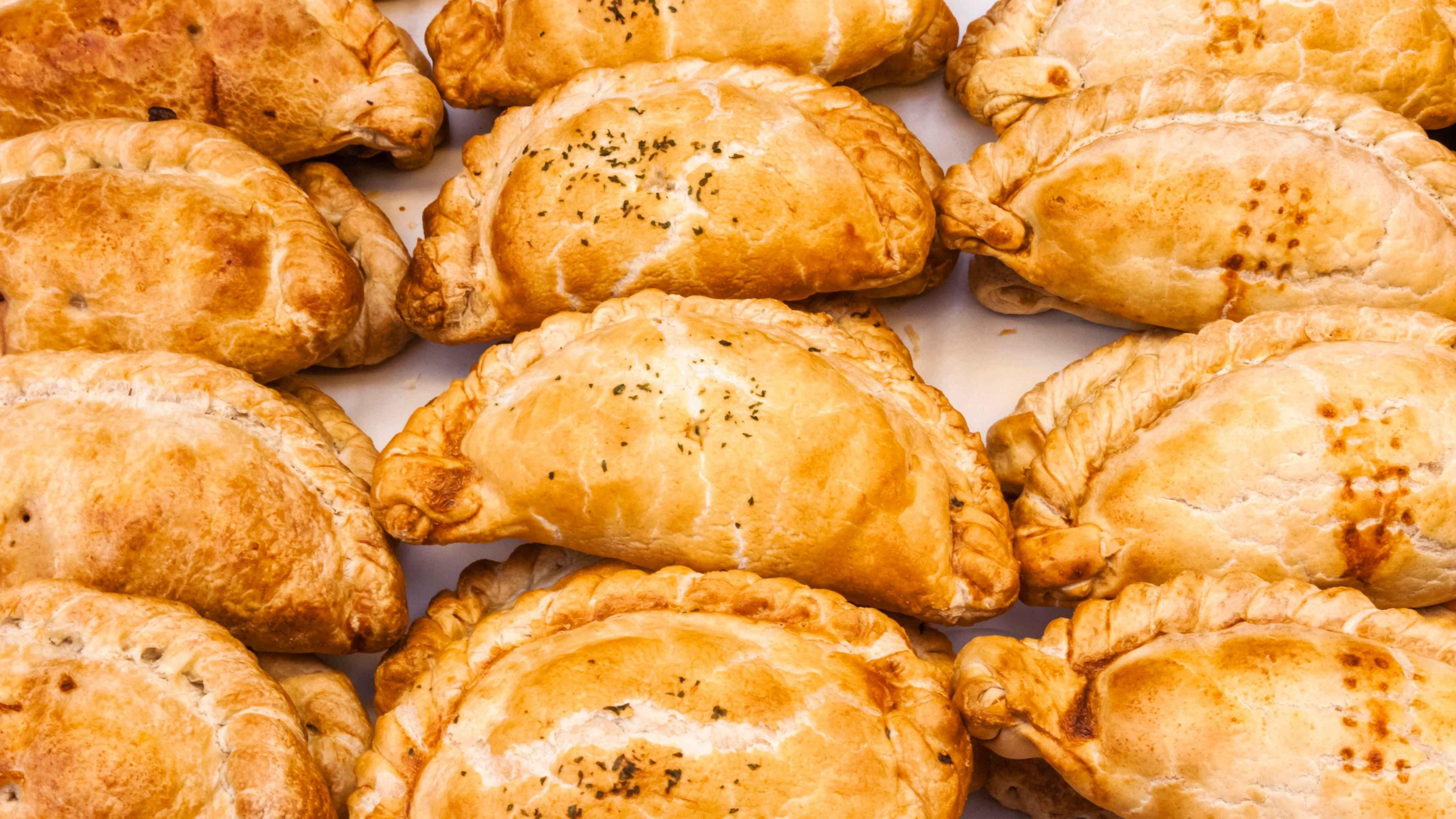 Twelve golden-coloured Cornish pasties are lined up in rows. They are standing on a white piece of paper.