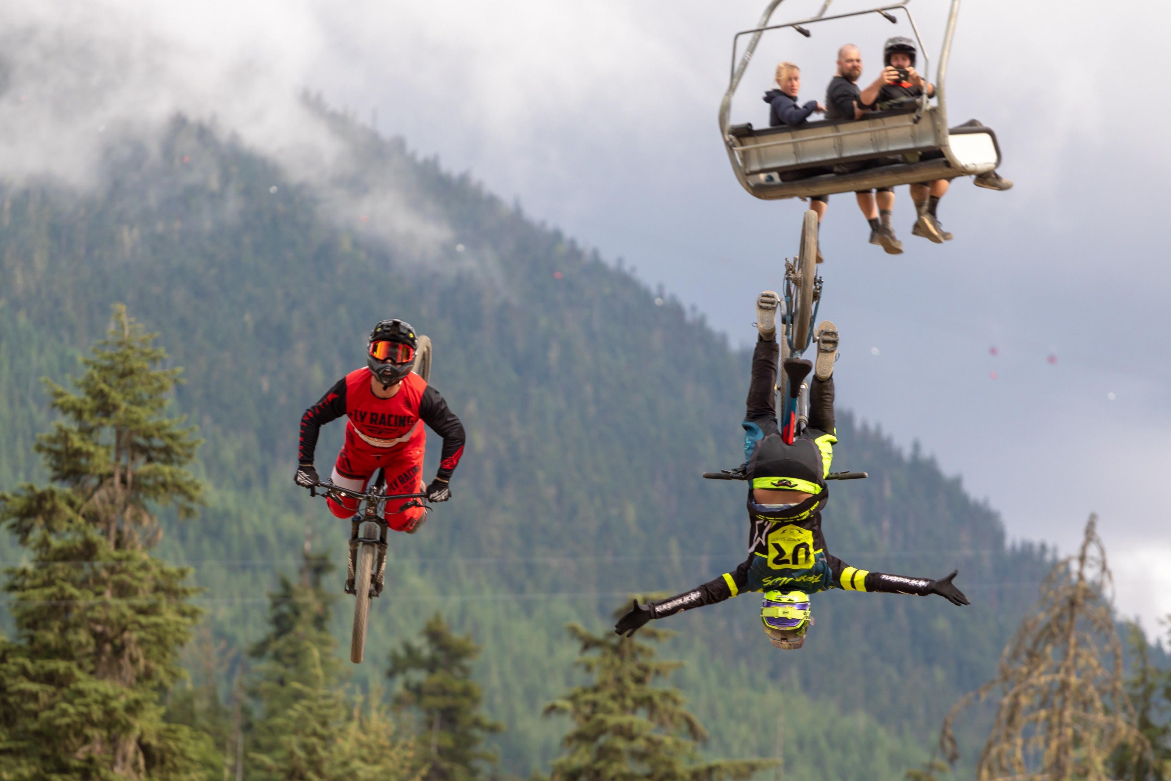 Two bikers in the air, one upside down with his arms out. Three people on a cable car watching from behind and taking a photo. 