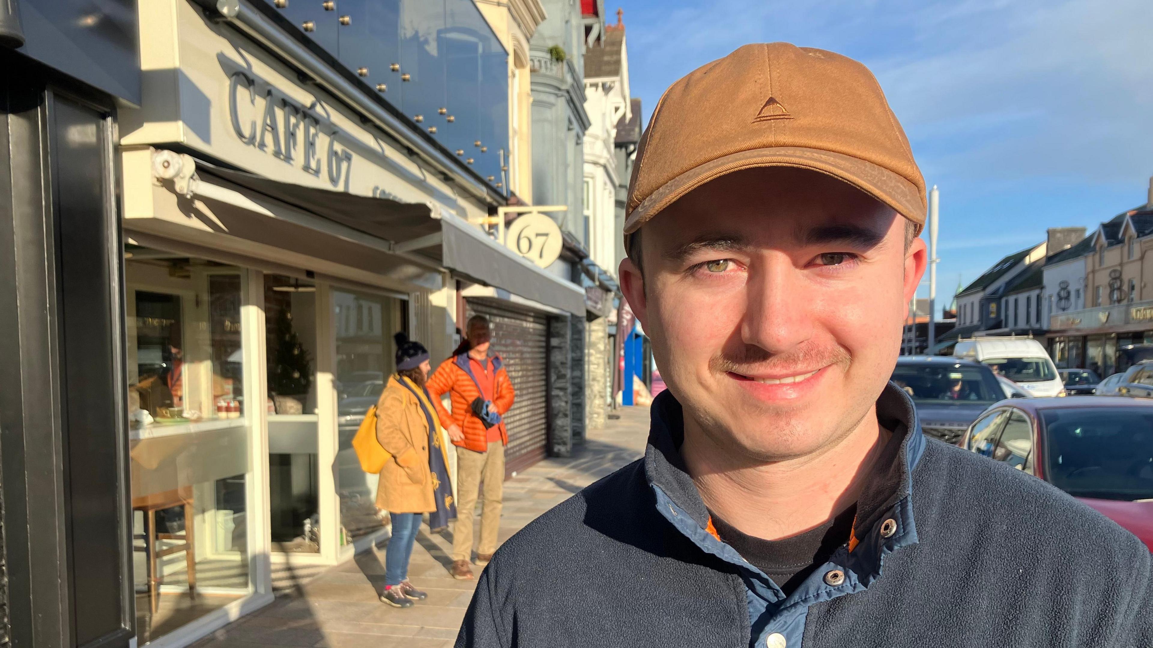 A man is smiling toward the camera. He is wearing a blue button-up jumper and a tan coloured hat. Behind him is a cafe on a busy street with people standing outside.