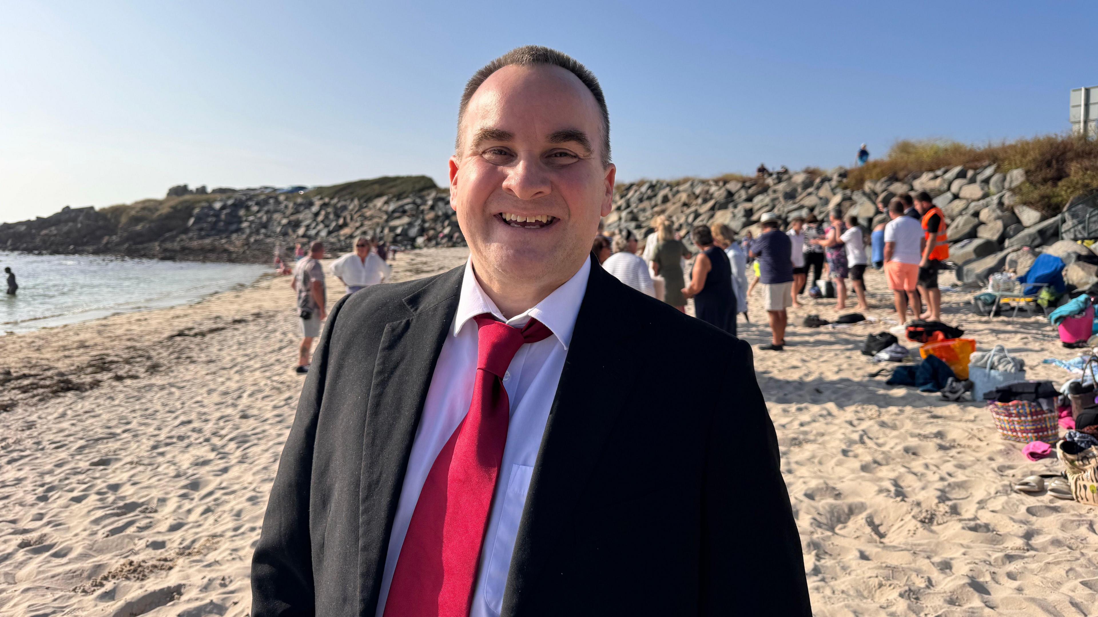 Tristan Boscher standing on a beach in office wear. Mr Boscher is wearing a suit and tie. Other swimmers are standing on the beach behind him. 