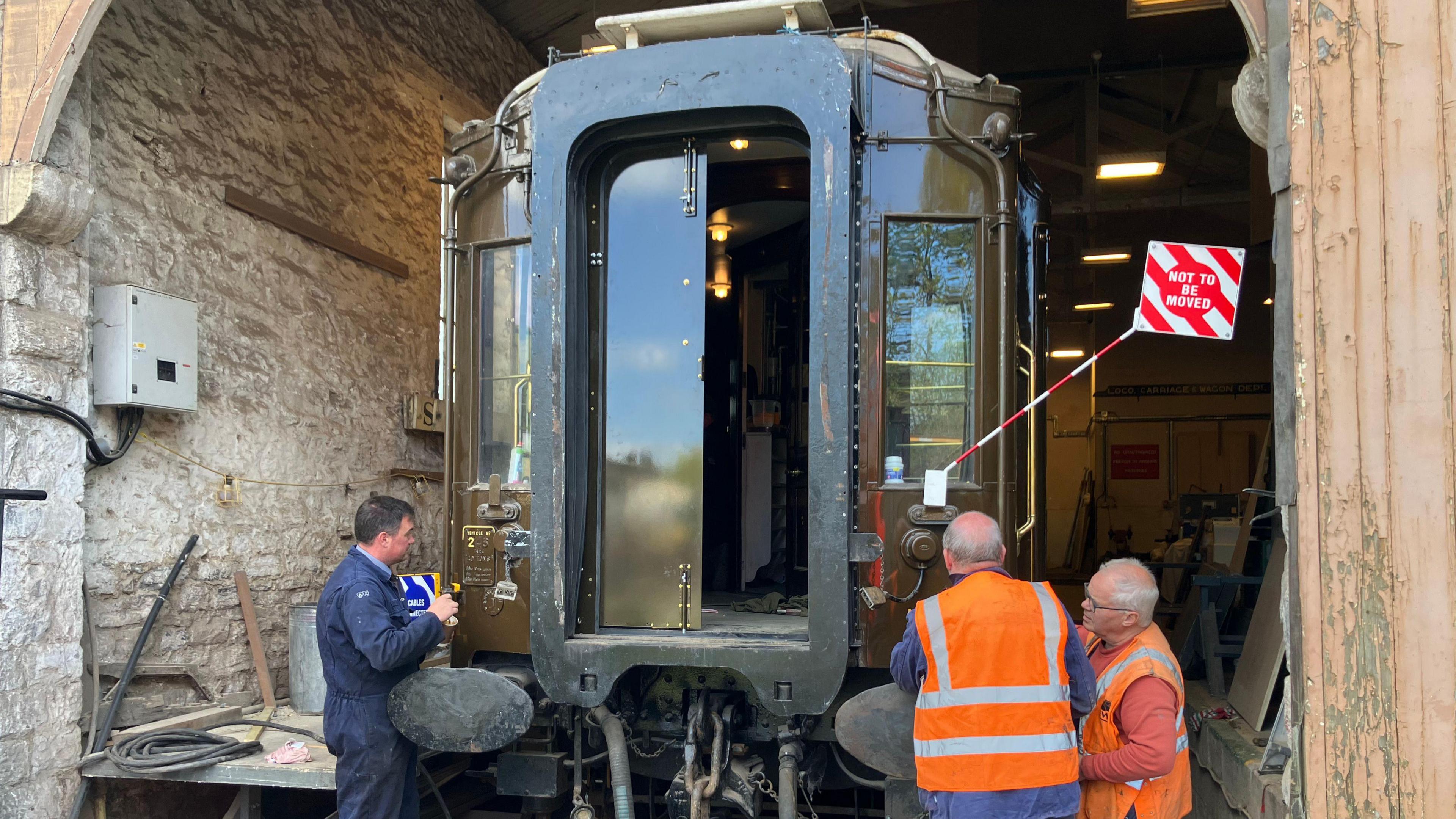 Three men working on the Pullman carriage. The outside of the carriage is brown. There is a "not to be moved" sign attached to the train. The man on the left is in blue overalls and the two men on the right are in orange high vis jackets. The wall of the workshop is a grey brick.