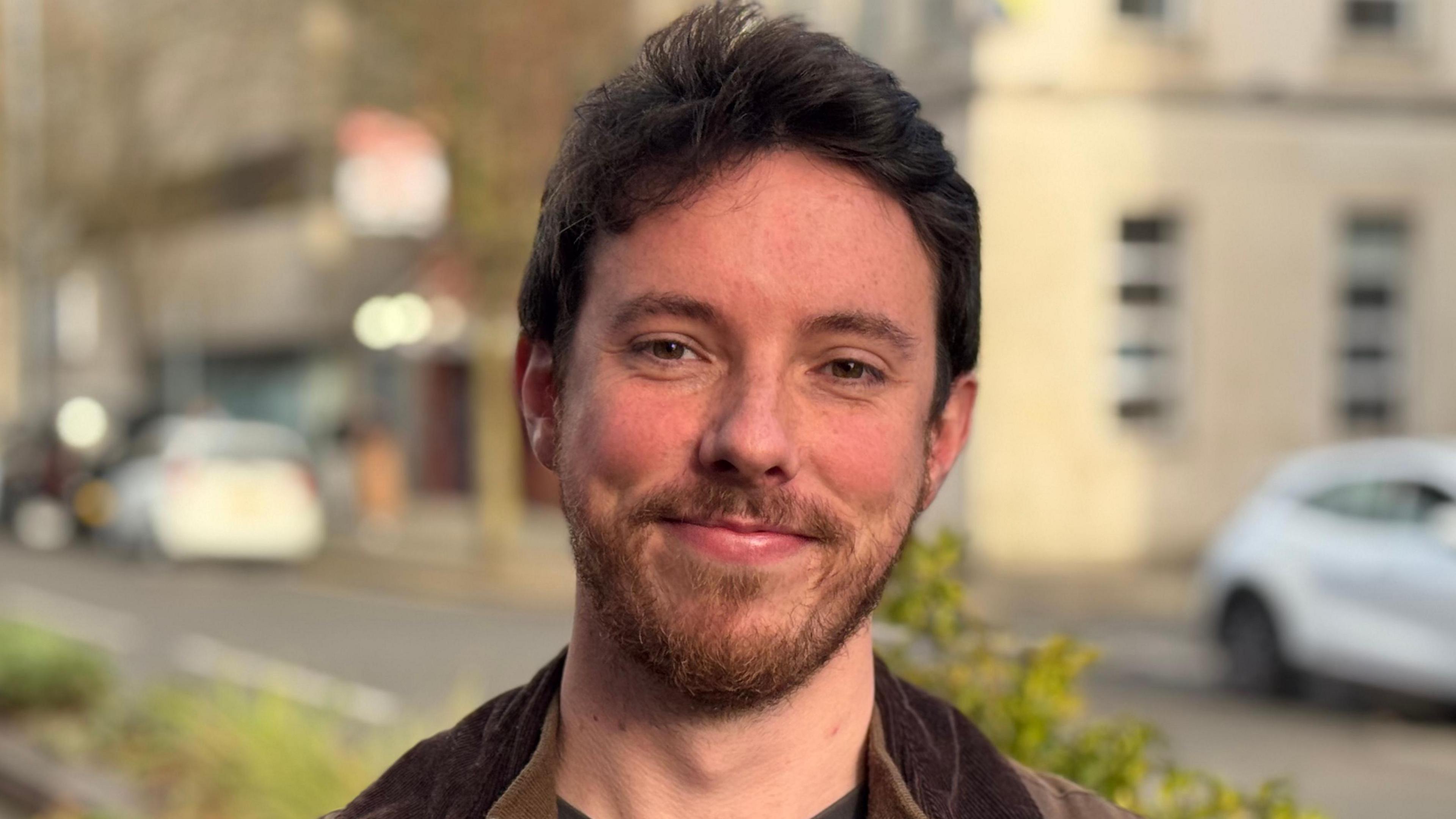 Shane Brady standing with a slight smile on his face. He is in his 20s, with black hair and a dark brown beard. He is wearing a dark brown wax jacket over a brown top and a dark grey t-shirt.