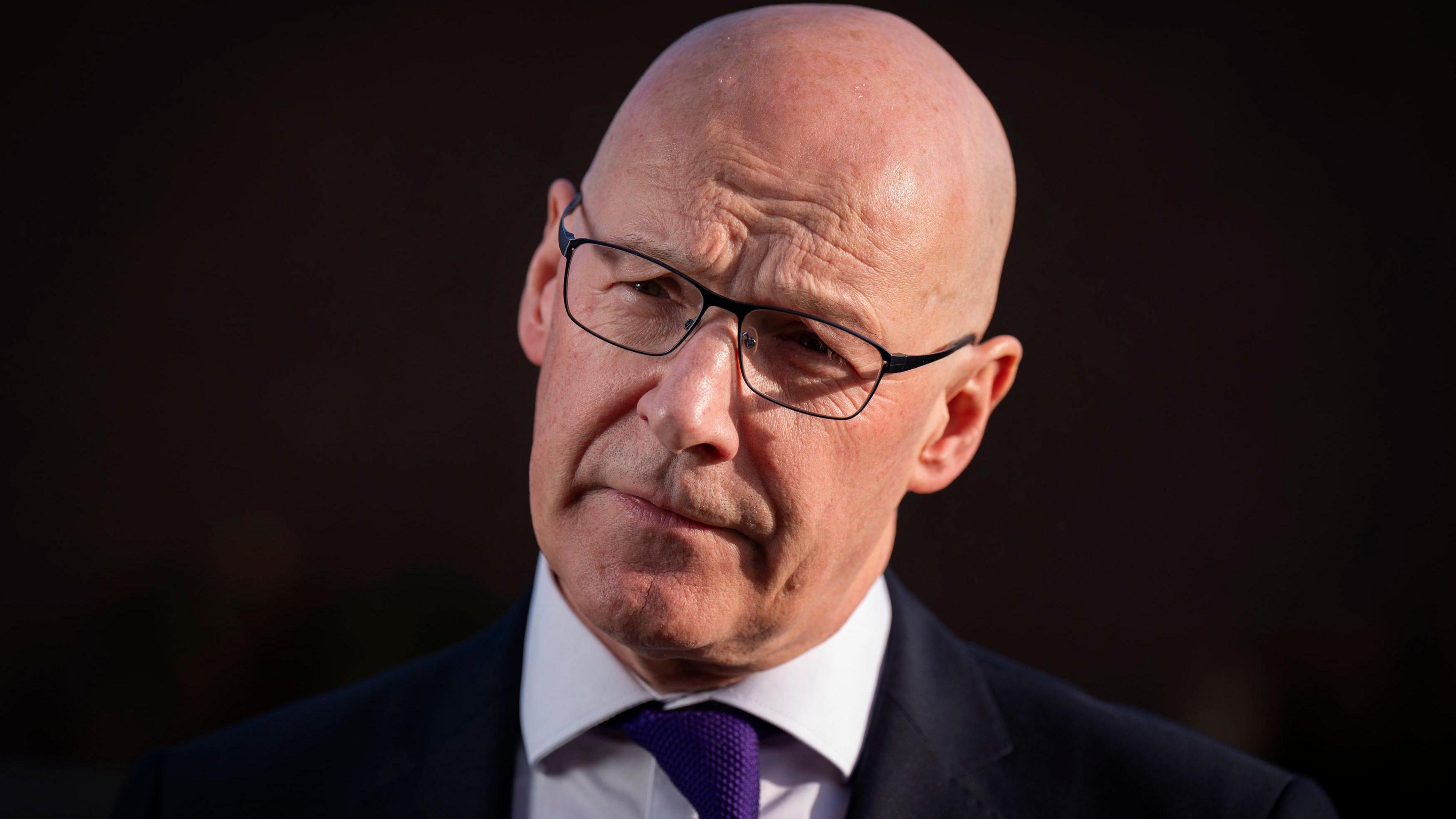 John Swinney, who has a bald head and is wearing glasses, stares off the left of the camera against a dark background. He is wearing a dark suit, white shirt and purple tie