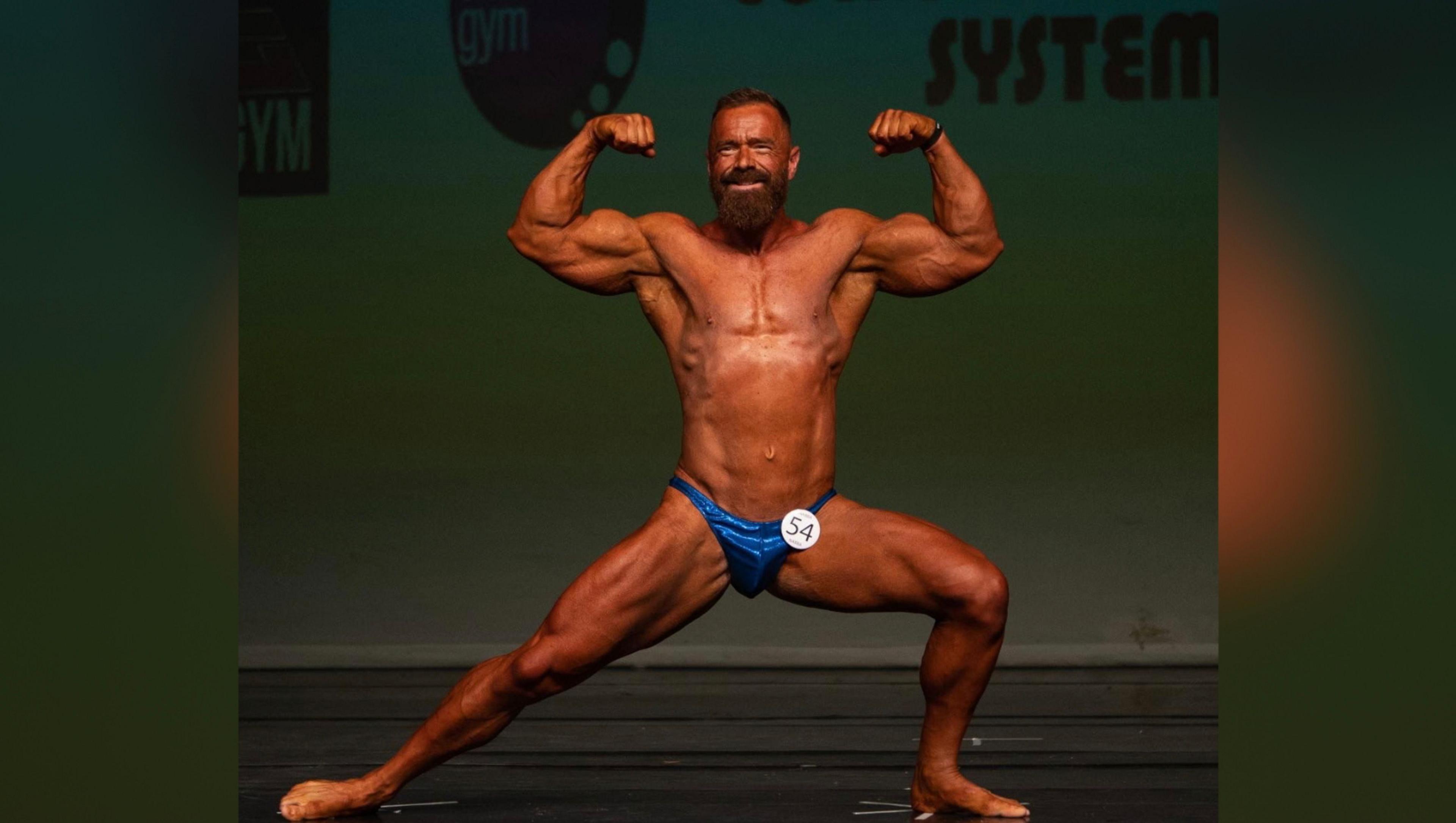 A man wearing small blue pants is seen with his arms in a muscle pose - he has defined muscles in his arms and stomach and legs has a tan.  He is taking part in a bodybuilding show and a badge with the number '54' can be seen pinned to his pants.