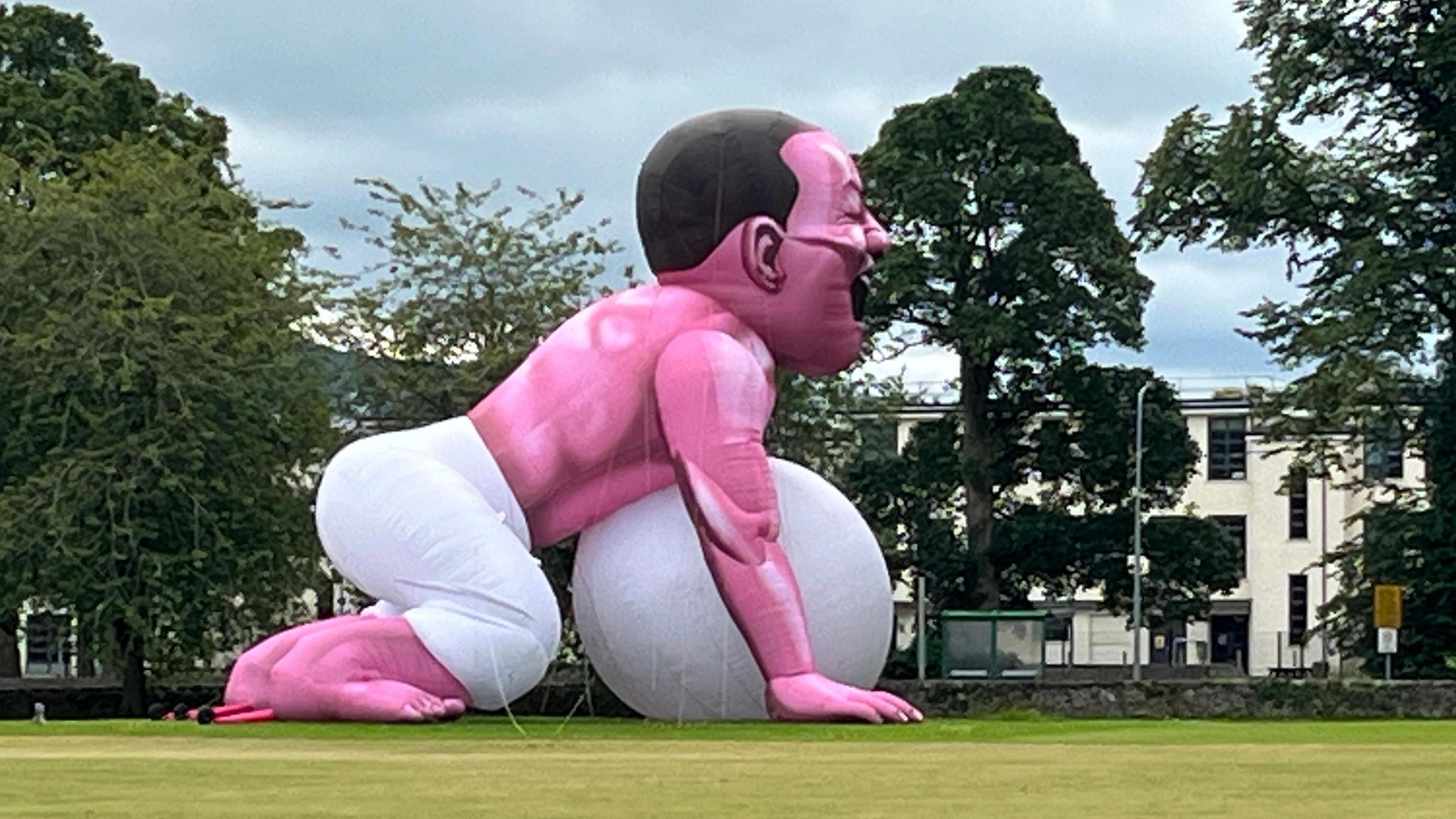 A side-on view of the giant inflatable topless man leaning over a giant white ball, placed in a school field