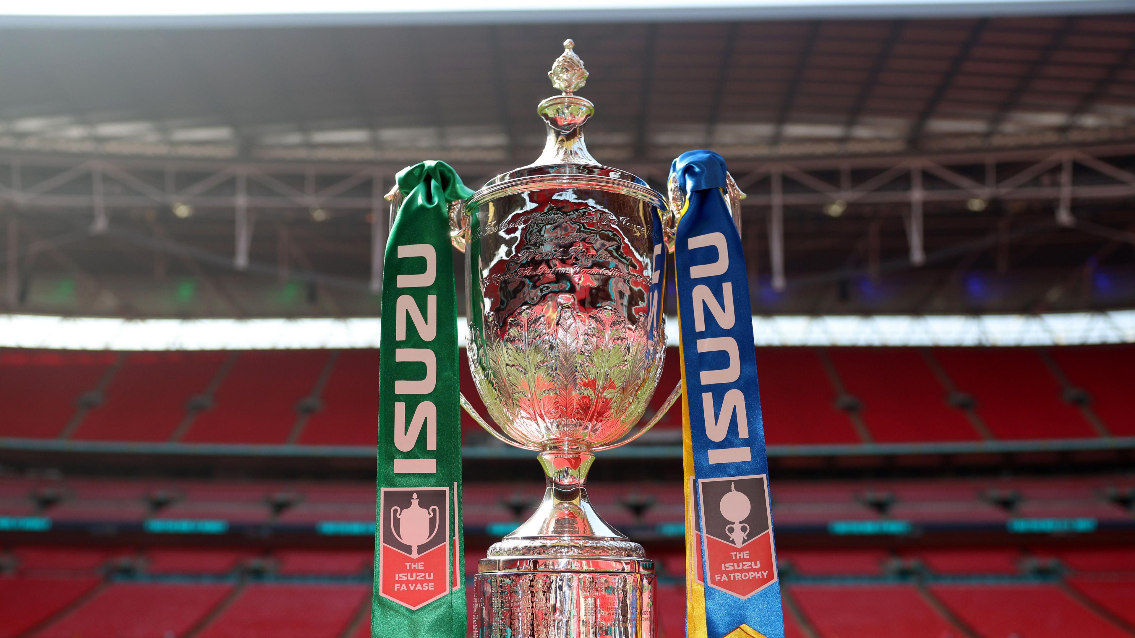 The silver trophy of the FA Vase displayed at Wembley Stadium, with empty red seats behind it and blue and green ribbons tied to each handle.