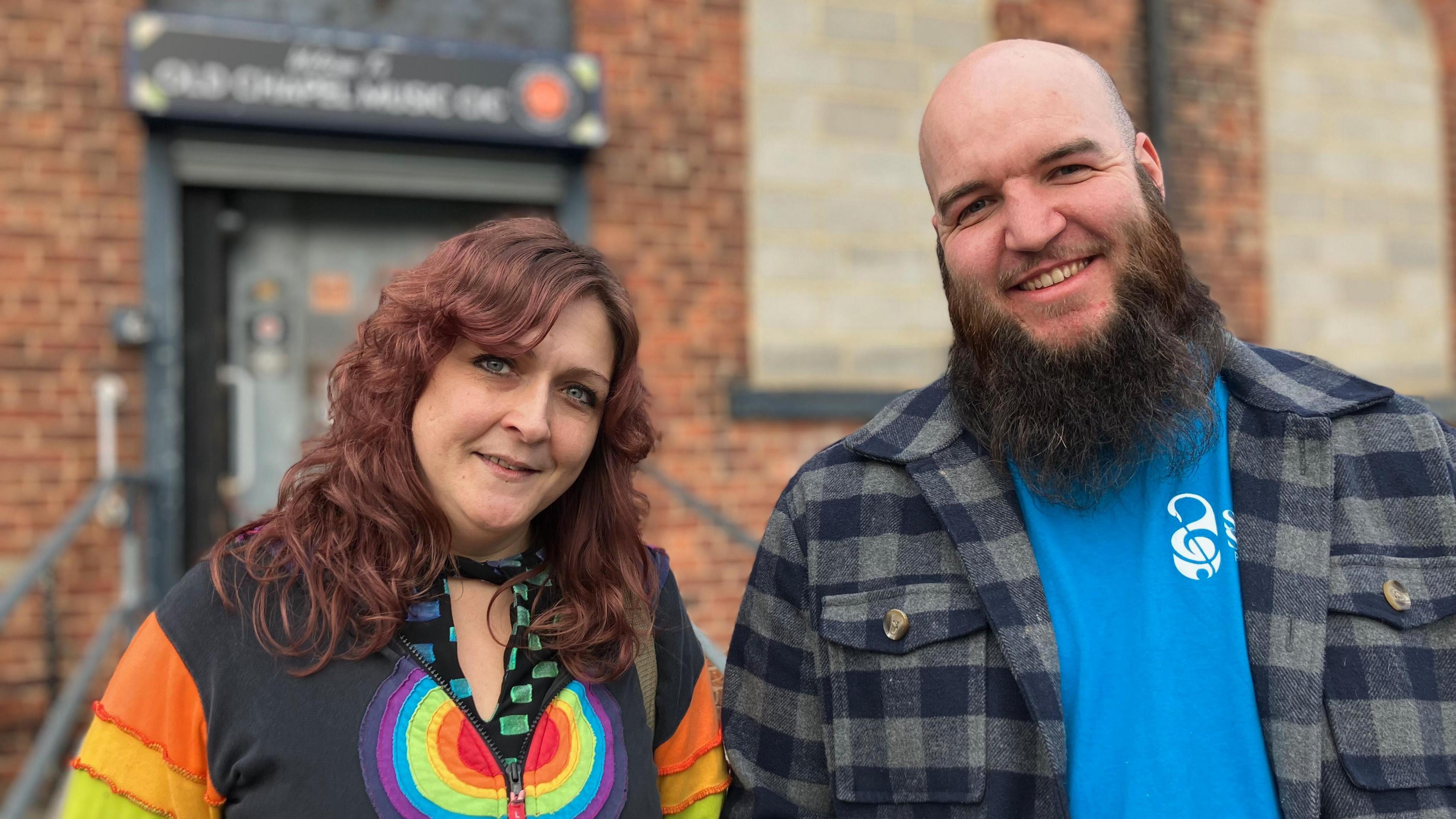 A tall man with a bald head and brown beard smiles broadly outside a redbrick building. He wears a navy checked shirt over a bright blue T-shirt, and stands alongside a woman with reddish-brown medium-length hair, who is wearing a colourful rainbow-striped cardigan.