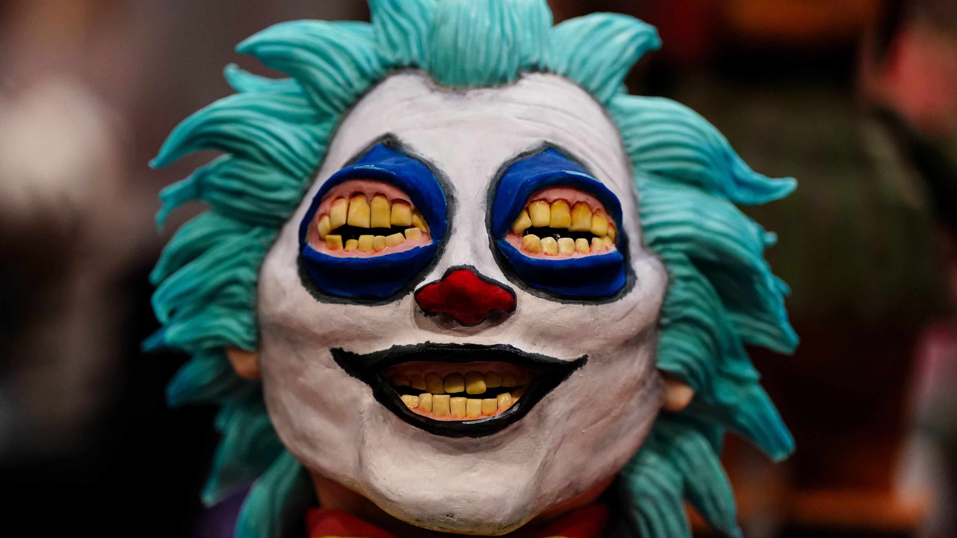 A close-up of a cake depicting a figure's head wearing clown make-up. He has spiky light blue hair and has teeth for eyes. He is grinning widely in a menacing fashion.