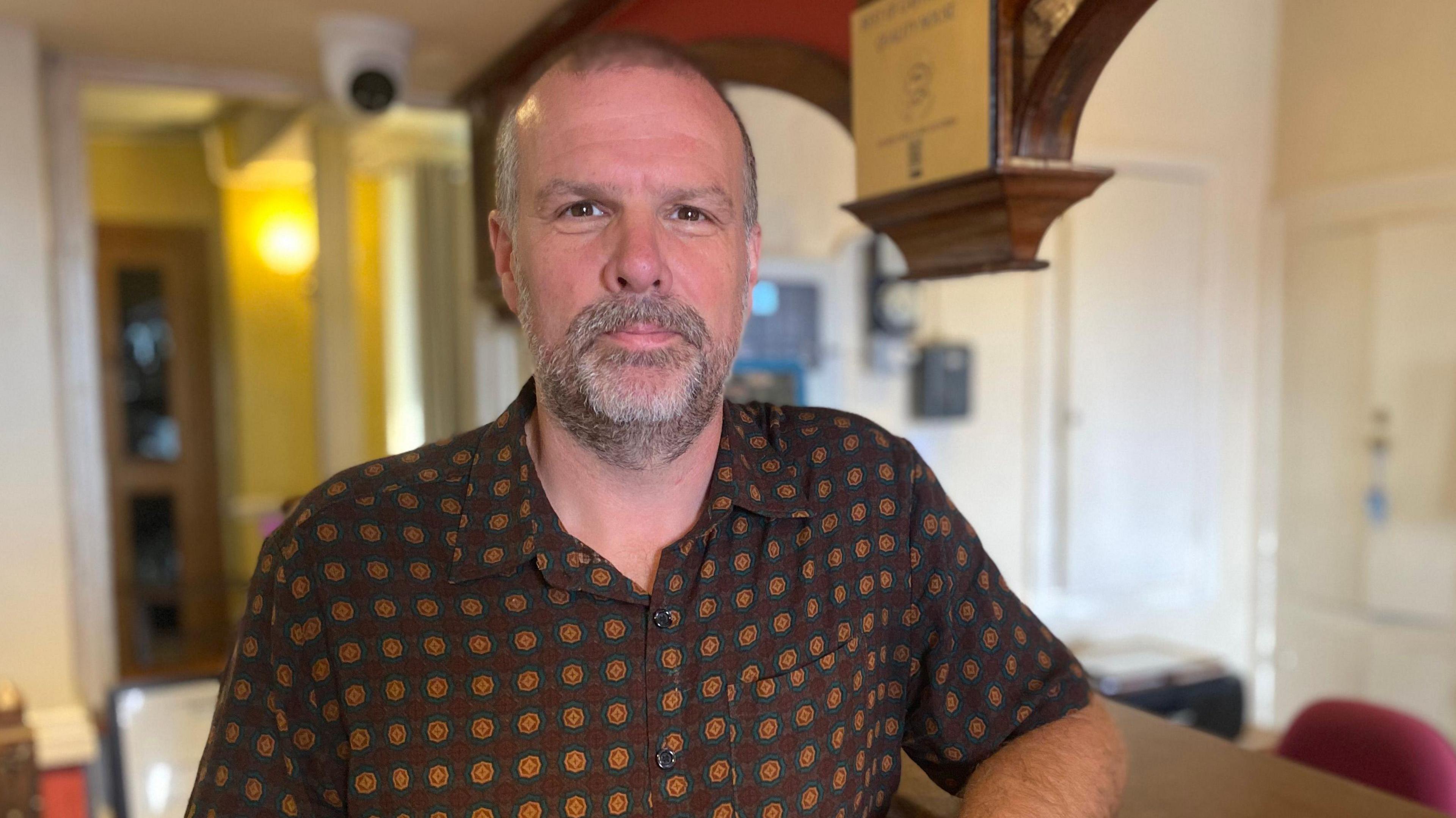 Huw is looking at the camera, he has a grey goatee and dark brown eyes. It is a head and shoulders shot of him stood inside a building which is blurred behind him. He is wearing a brown shirt with a dark yellow and green circular pattern on it.
