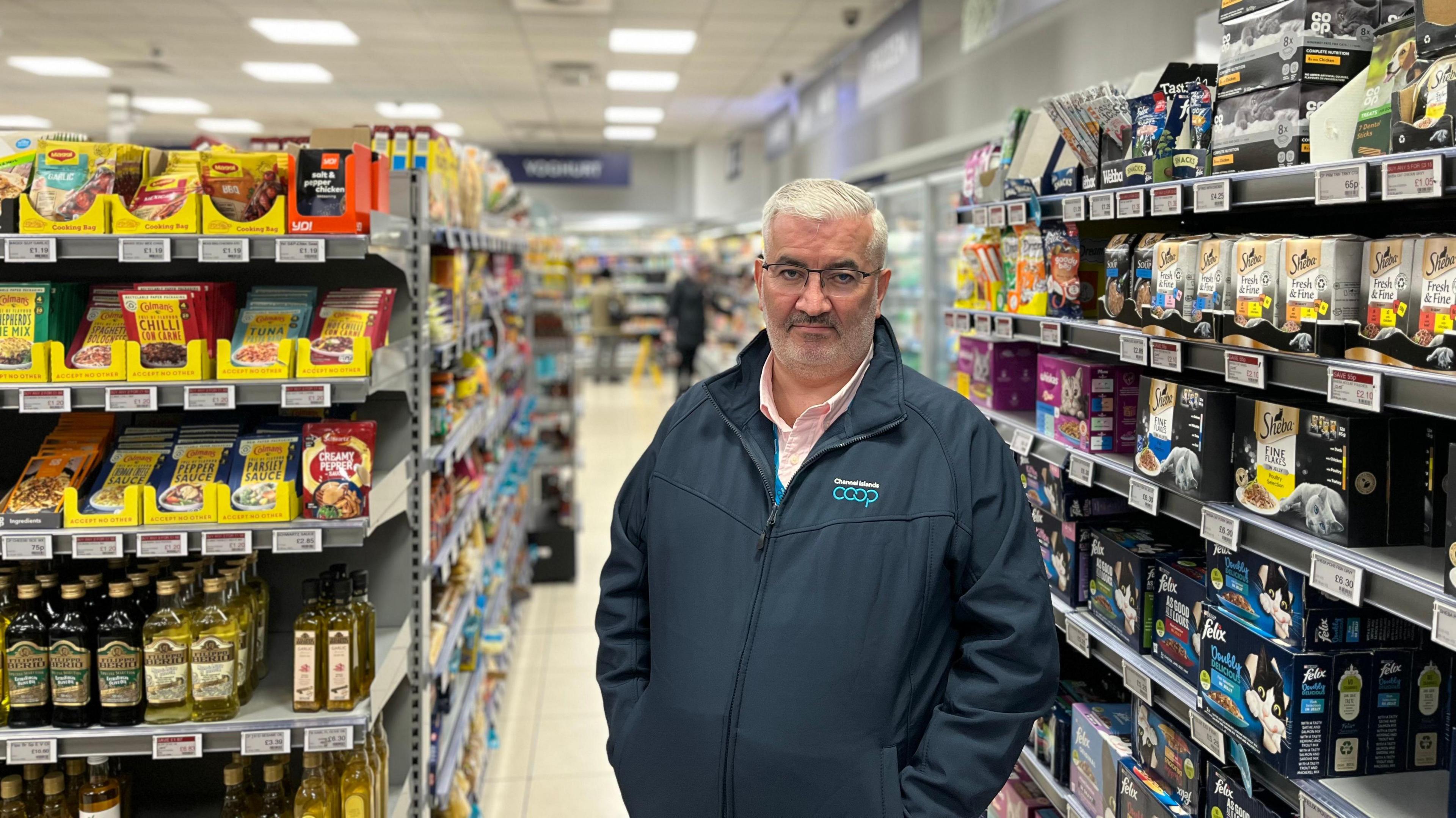 Mark Cox, a man with white hair, glasses and a grey beard. He is standing in a supermarket aisle, with pet food to the right of the image and cooking supplies - including olive oil and seasoning - to the left. He is wearing a 'coop' branded jacket with a zip through the middle, over a pink shirt.