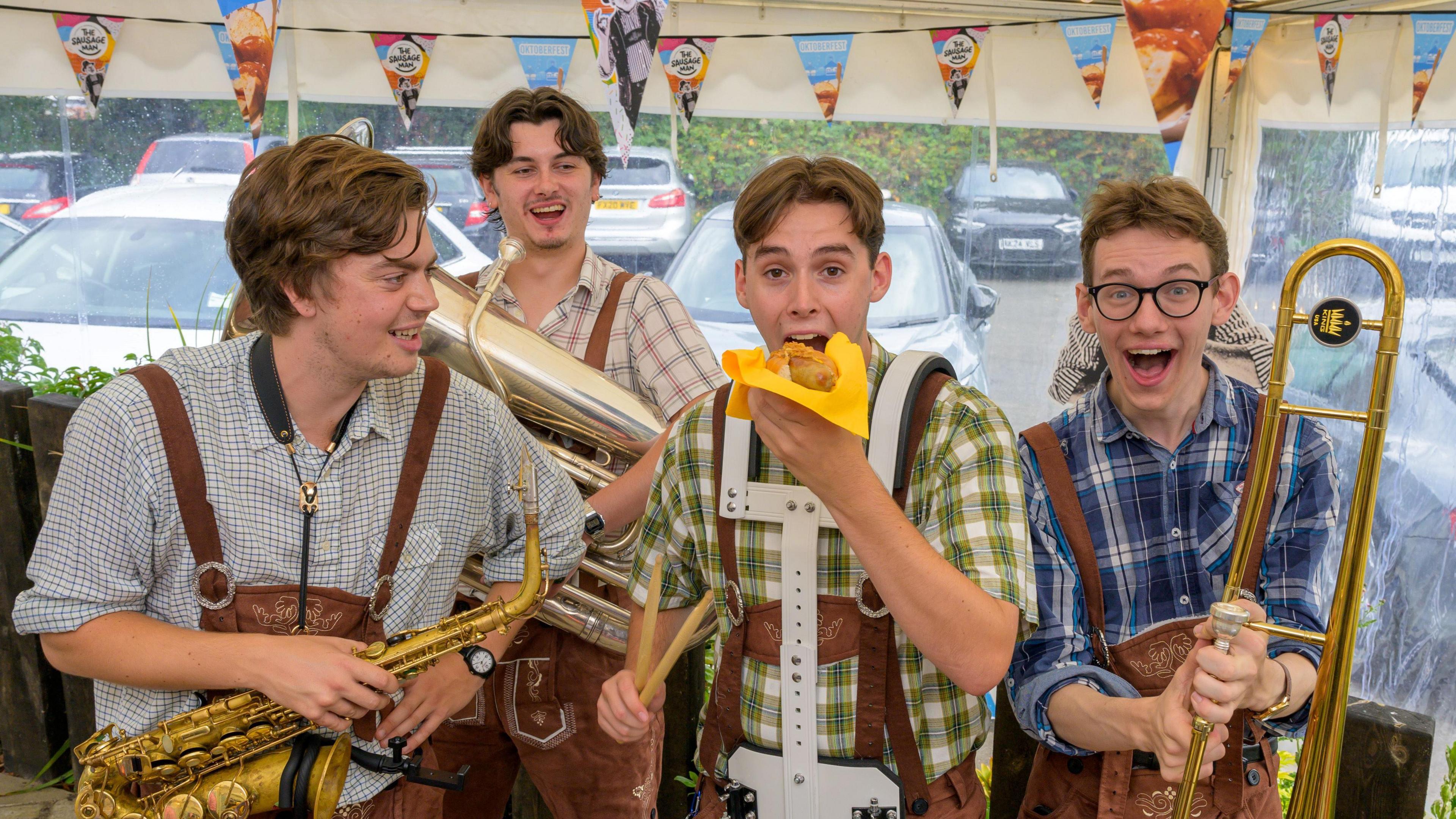 Four members of a band, holding musical instruments, with one band member seemingly about to eat a hot dog. They appear to be in a gazebo with clear sides, looking out onto a car park.