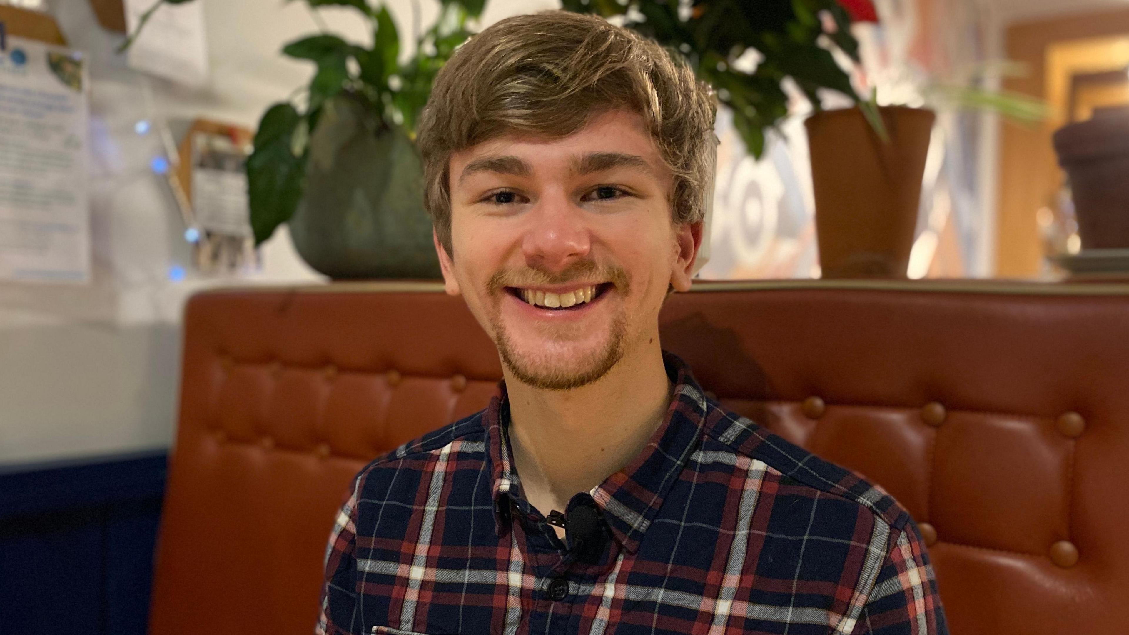 Sam Braham has short brown hair and is wearing a checkered shirt. He is sitting on a brown sofa 