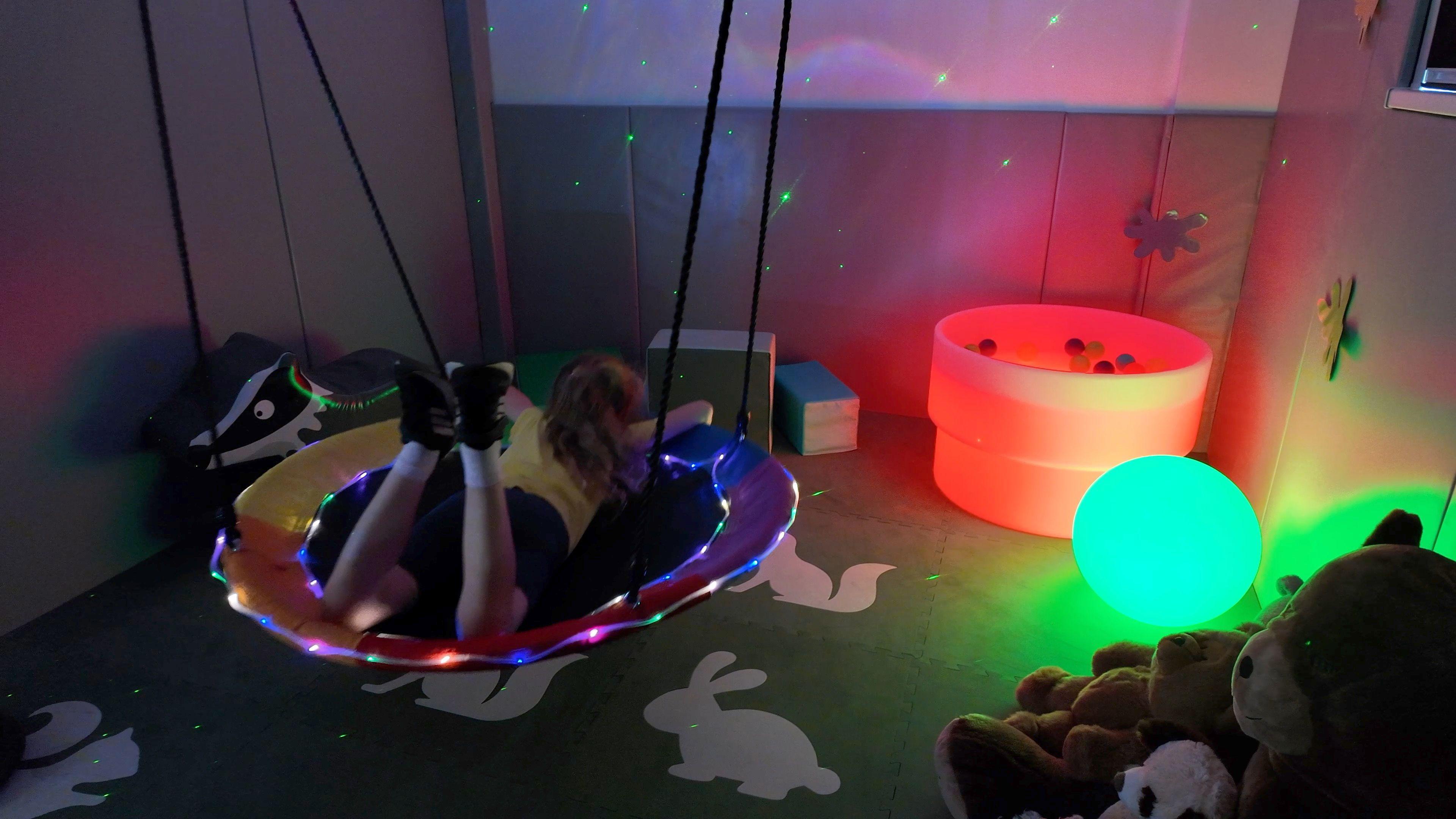 Darkened room with a lit up swing and some coloured balls and a pit in the right hand corner. The floor is covered in soft tiles with animals on and the walls are covered in soft padding.