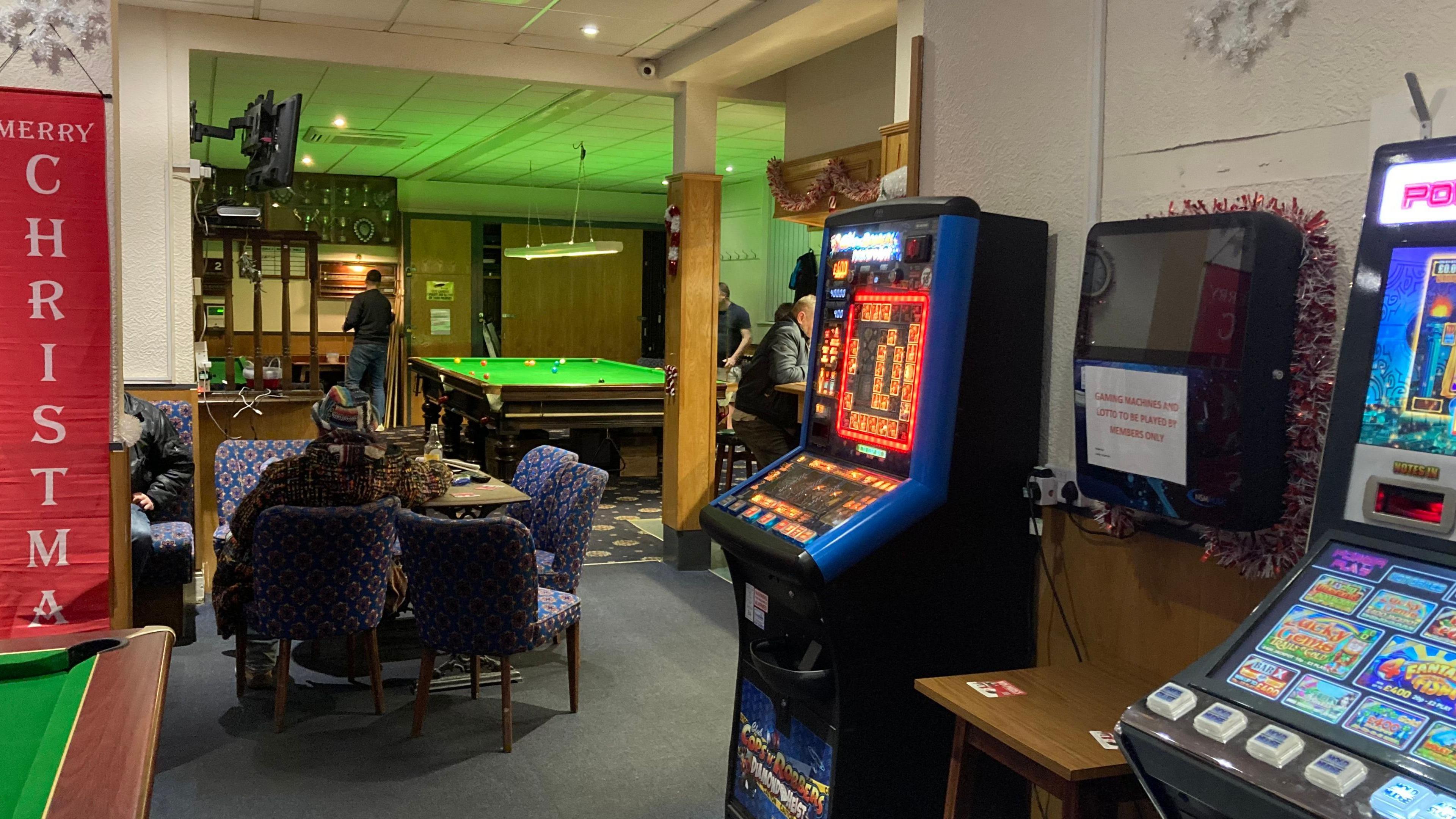 A view across a social club with the corner of a snooker table to the left and slot machines to the right. A red banner has white text reading "Merry Christmas". In the middle-distance a woman sits in a comfortable bar chair at a table. In the background a man can be seen standing next to a snooker table. 