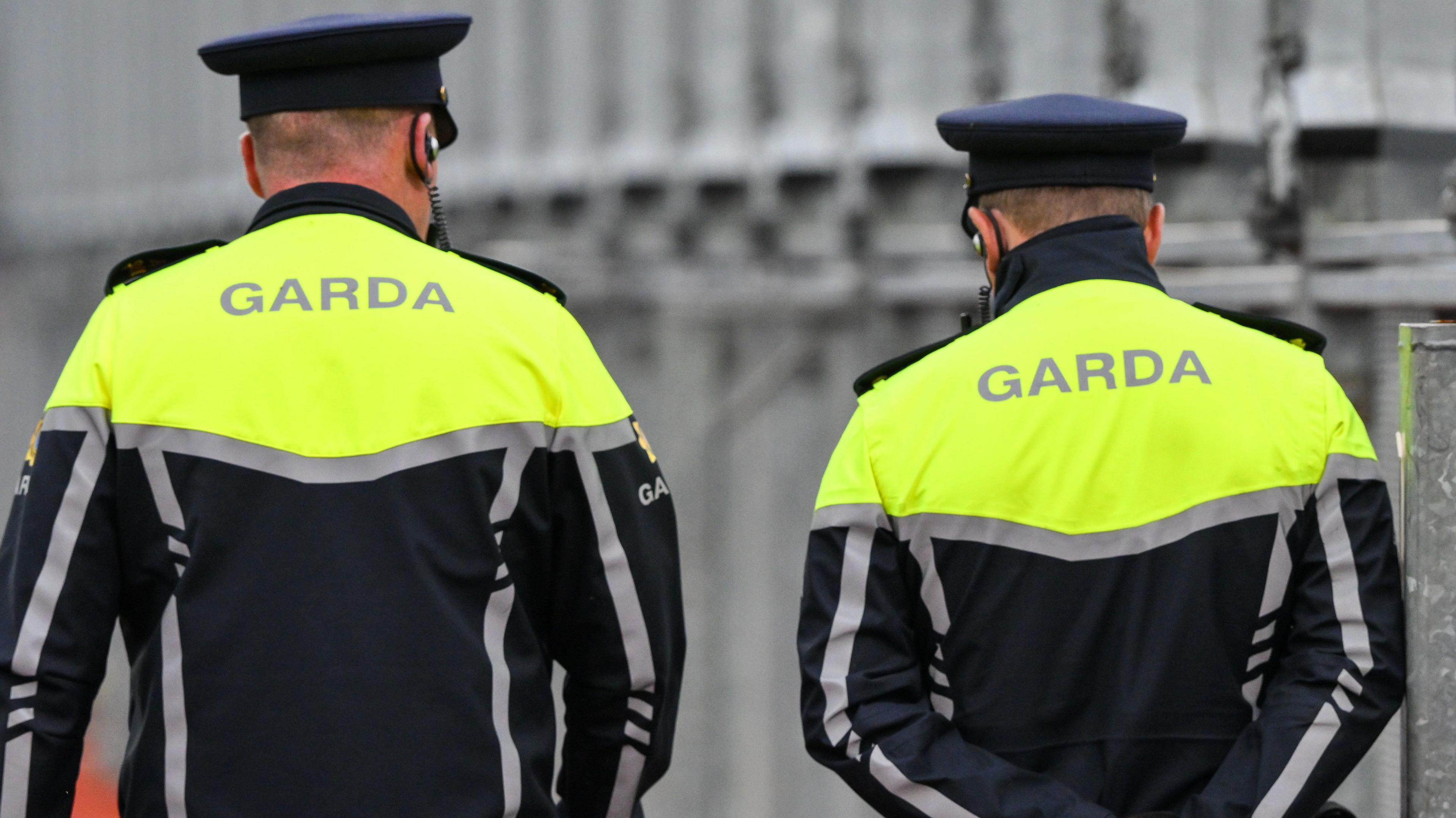 The backs of two gardaí officers in their uniforms. They are both wearing flat-top hats, ear pieces and a reflective jacket with 'gardaí' on the back.