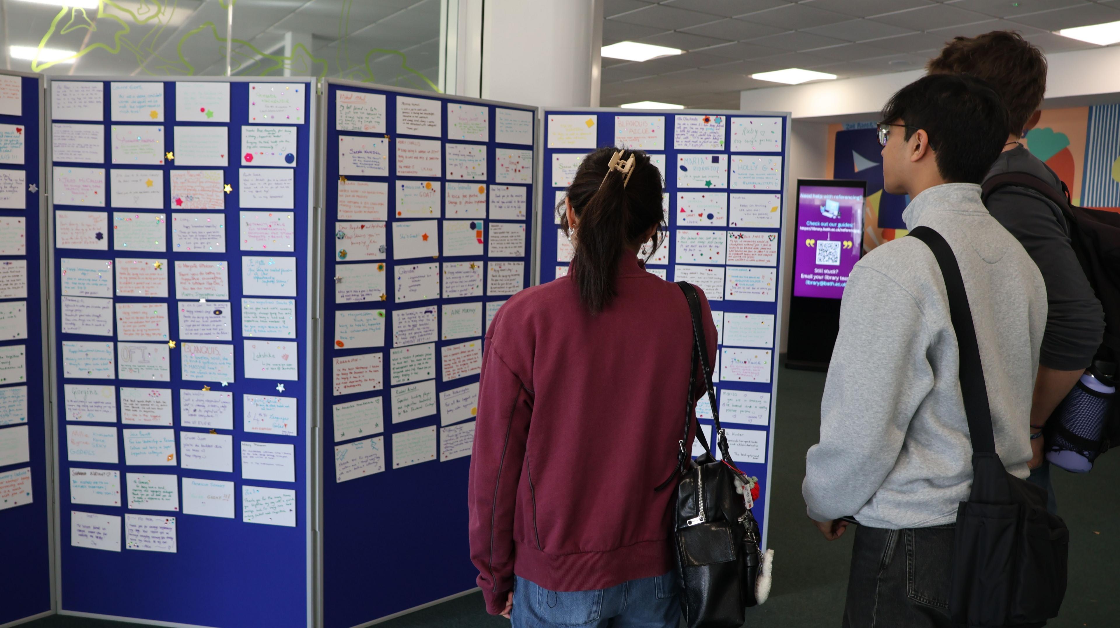 A woman wearing a burgundy sweatshirt and blue jeans, with her black hair up in a claw clip. Beside her are two men, one wearing a grey sweatshirt with a black cross body bag and the other wearing a dark-grey shirt. They are reading the wall of nominations which are pinned to a blue display board.