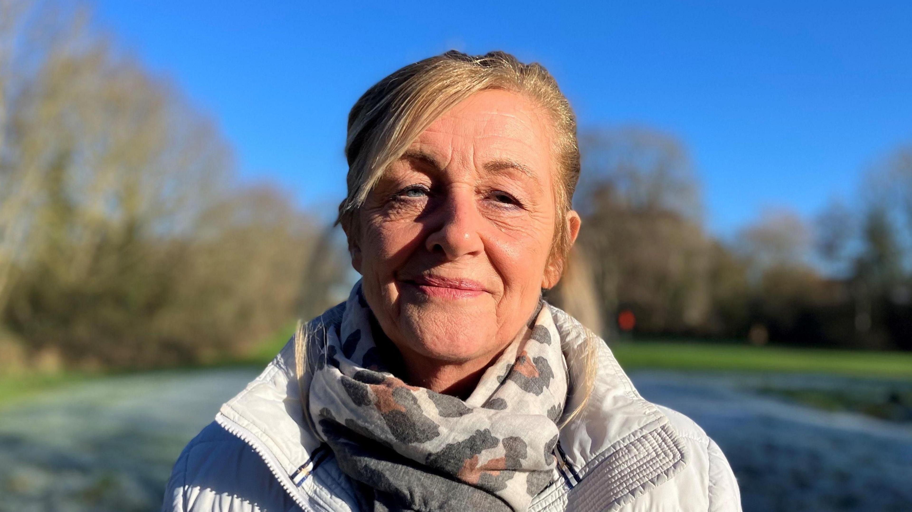 A woman with fair hair, wearing a patterned scarf and white coat, looks at the camera. She is standing against a blurred rural background with a blue sky and frost on the grass. 