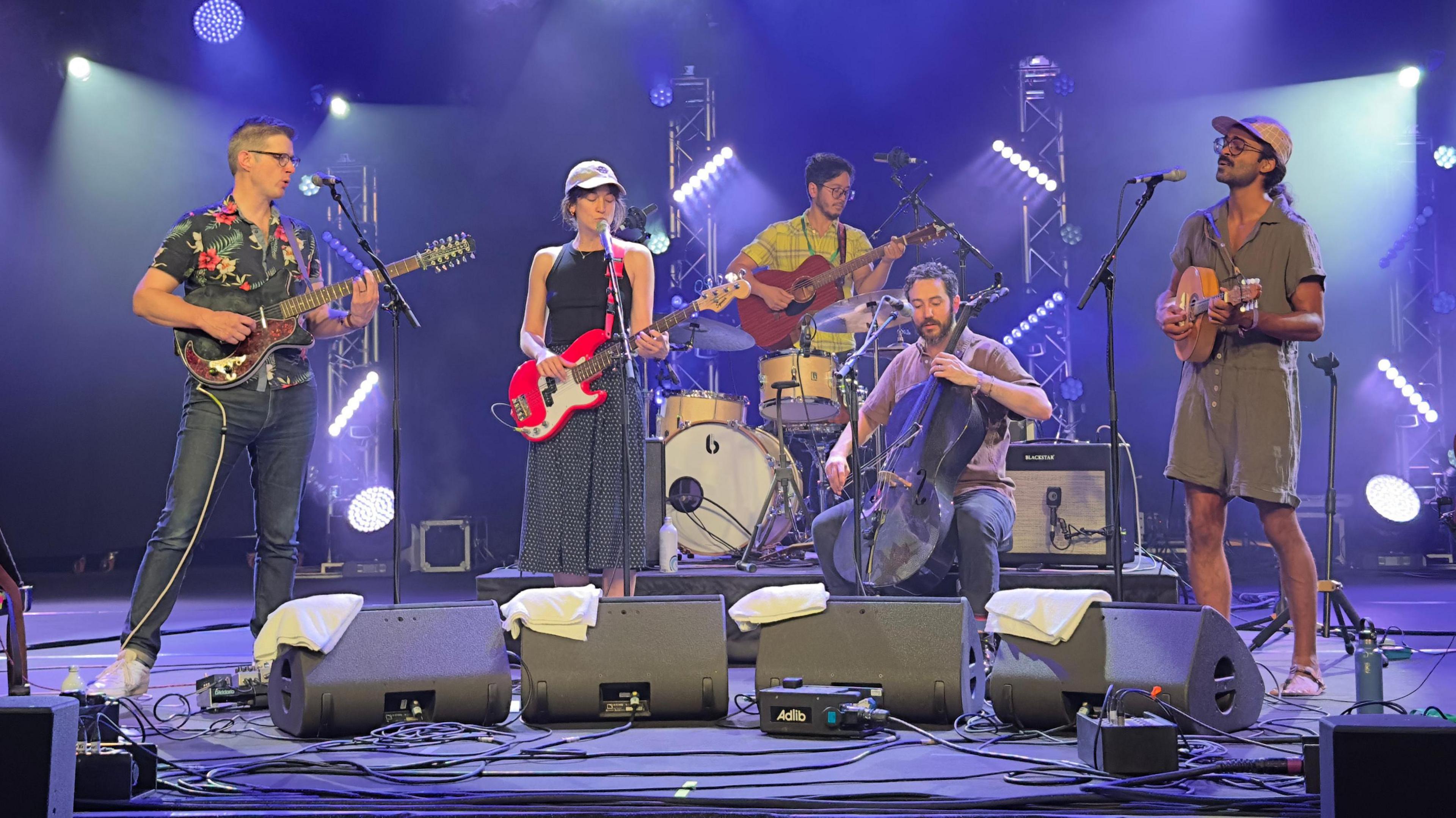 Five members of a folk band - four men and a woman are on a stage performing. They are playing drums, guitars, double bass and a ukulele. 