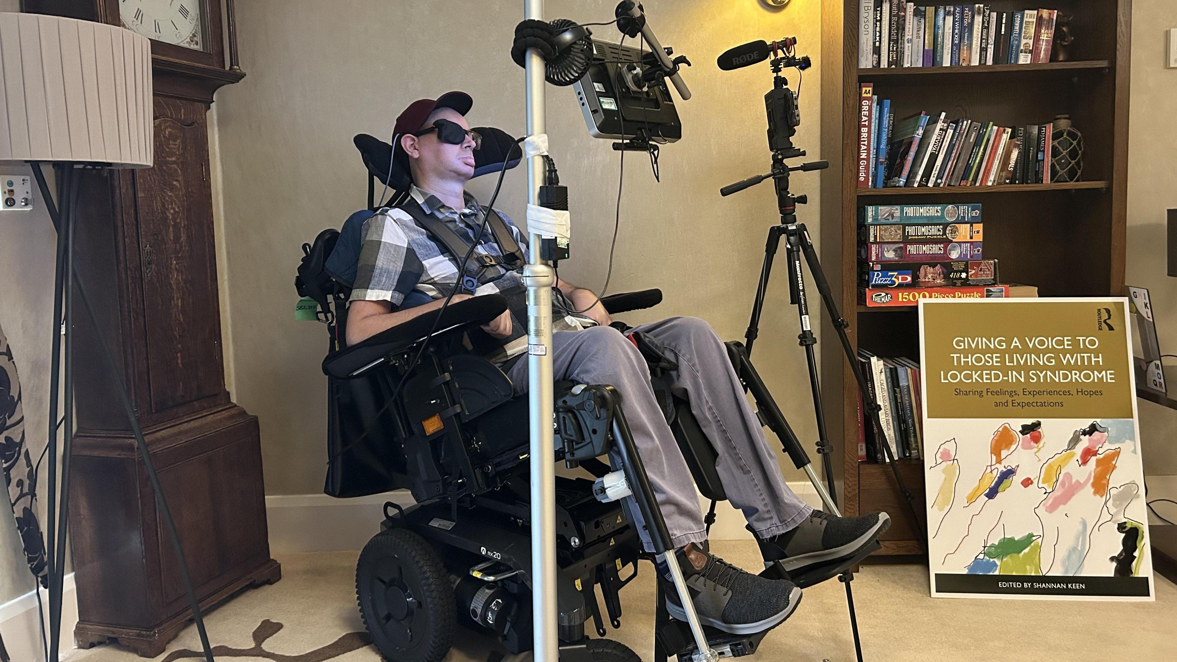 Bram Harrison in his wheelchair in a room in the care home he lives in in Exeter. Behind him a grandfather clock. He is in front of a computer screen and there is a poster about the book he has contributed to.