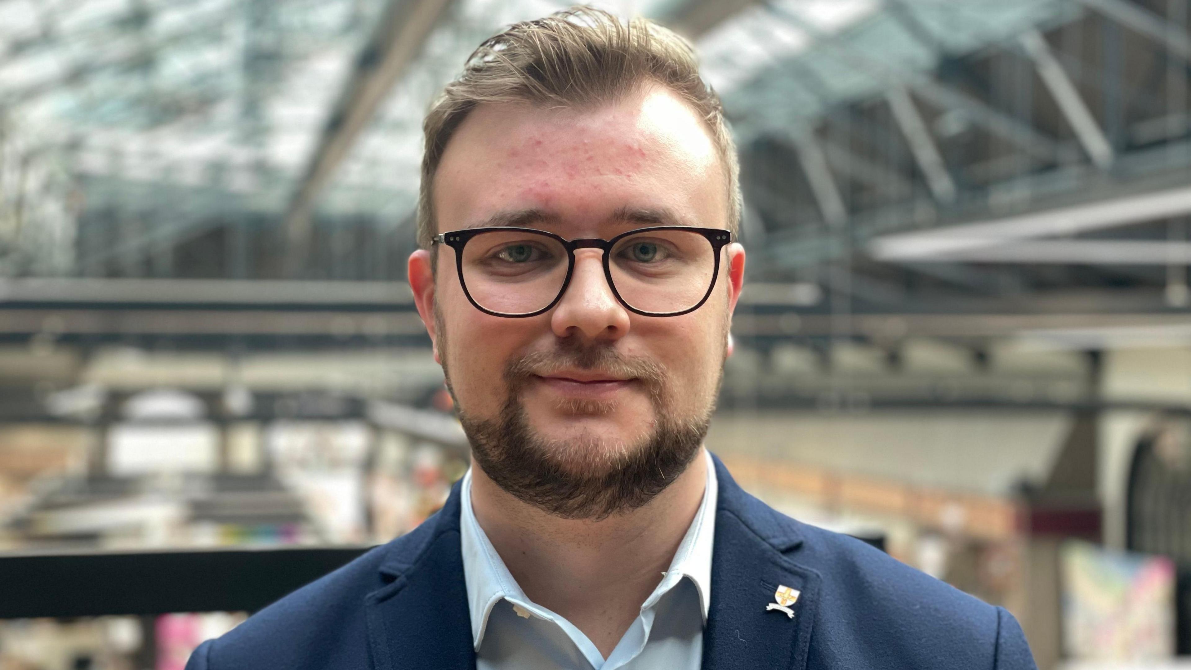 Joshua Wells stares into the camera with the cornhill market behind him. He is smiling and wearing a blue suit and light blue shirt. He has a beard, brown hair and dark rimmed glasses.