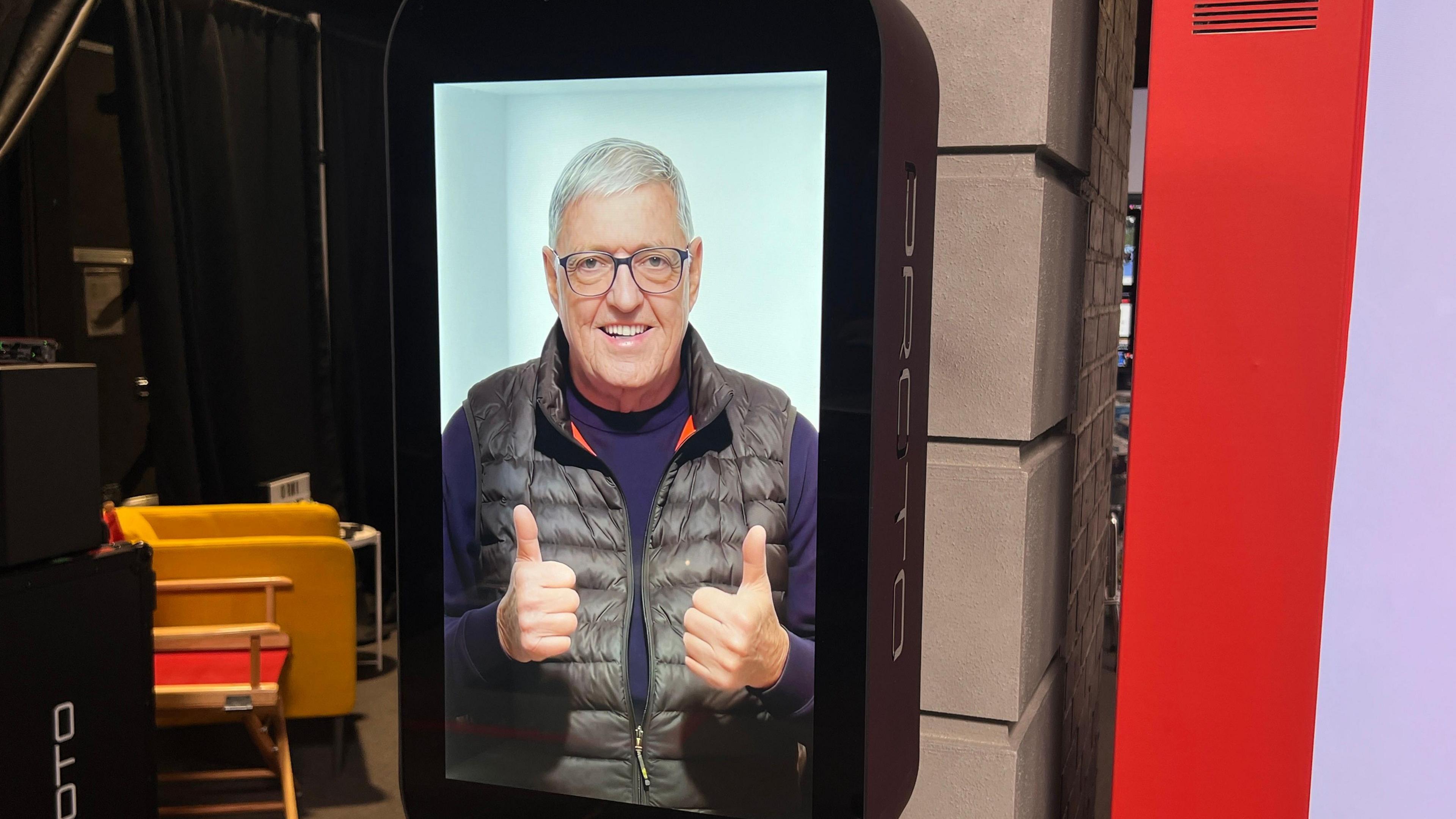 Picture of hologram version of Bill on a large screen, wearing a purple jumper and black gilet. He smiles and gives two thumbs up.