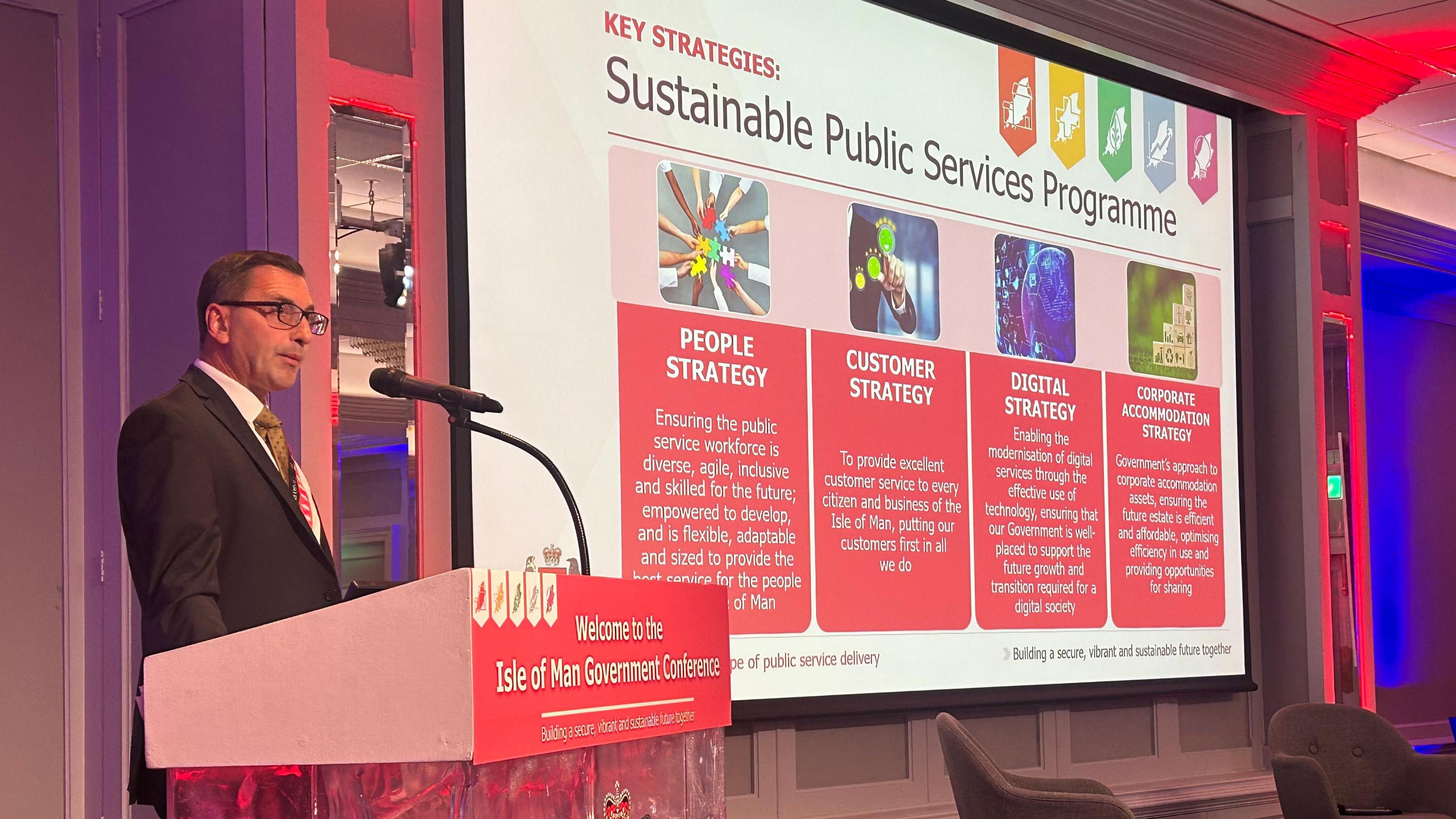 Andy Ralphs standing at a lectern in front of a microphone. He is wearing a dark suit, white shirt and yellow tie with a large digital screen to his side outlining the four strands of the Sustainable Public Services Programme.