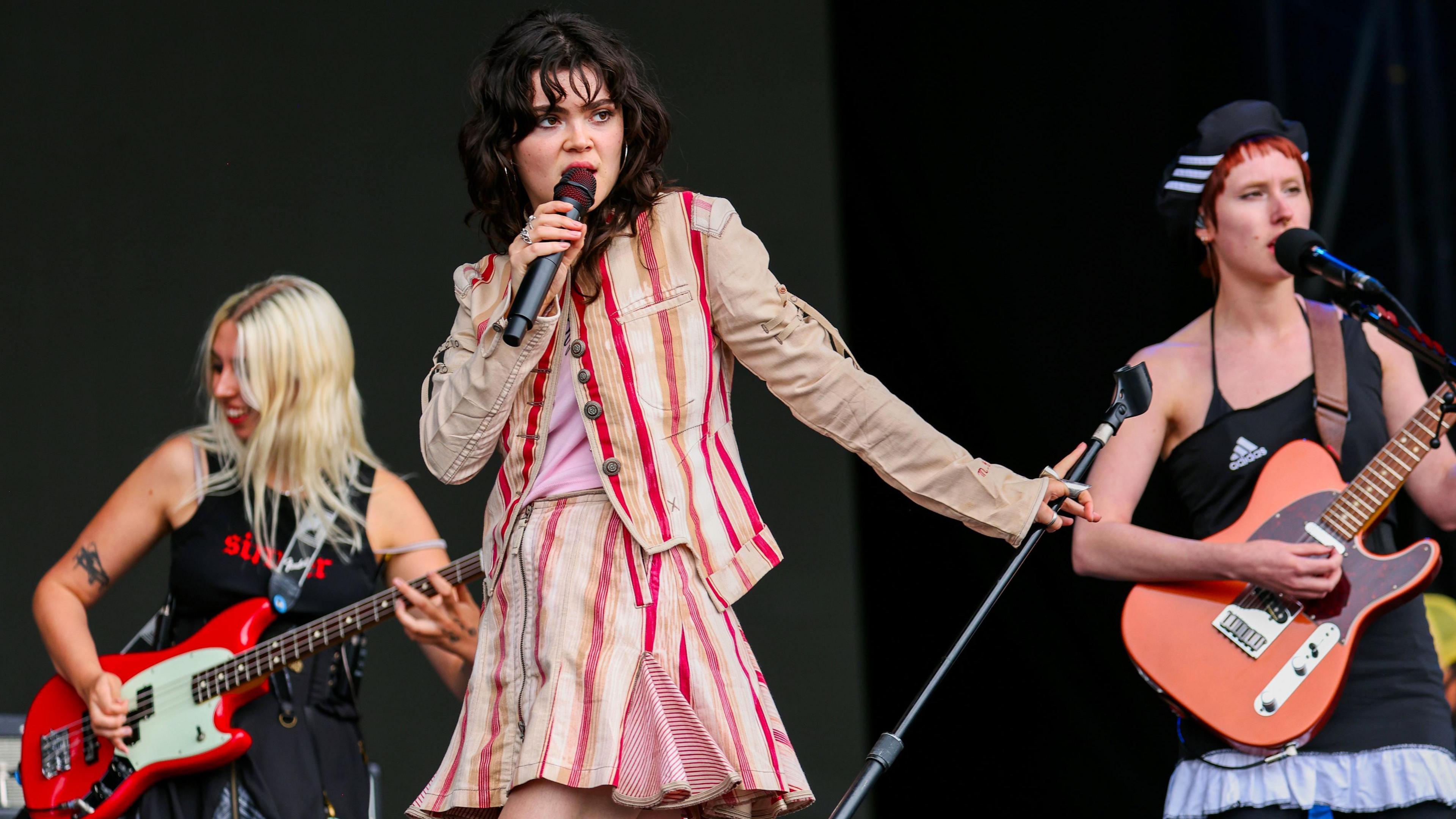 The Last Dinner Party performing on stage at Reading Festival. Lead singer Abagail wears a red and white-striped two piece suit and stands in front of her bandmates on guitar. 