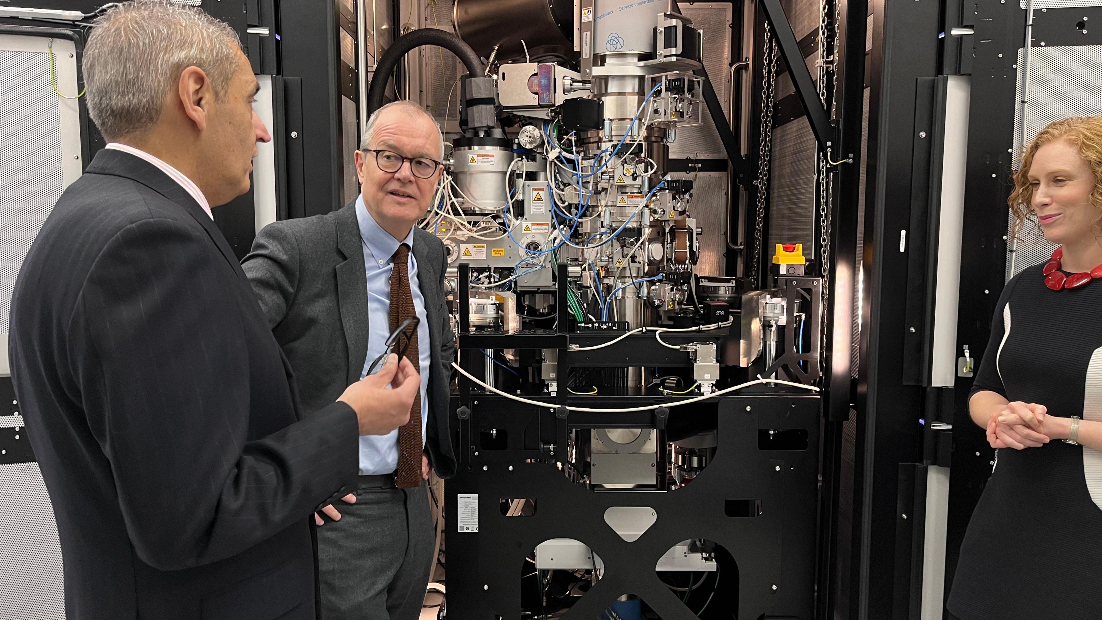 Patrick Vallance listening to a man speaking. A woman is standing to the side of a large bit of machinery with the two men on the other side of it. 