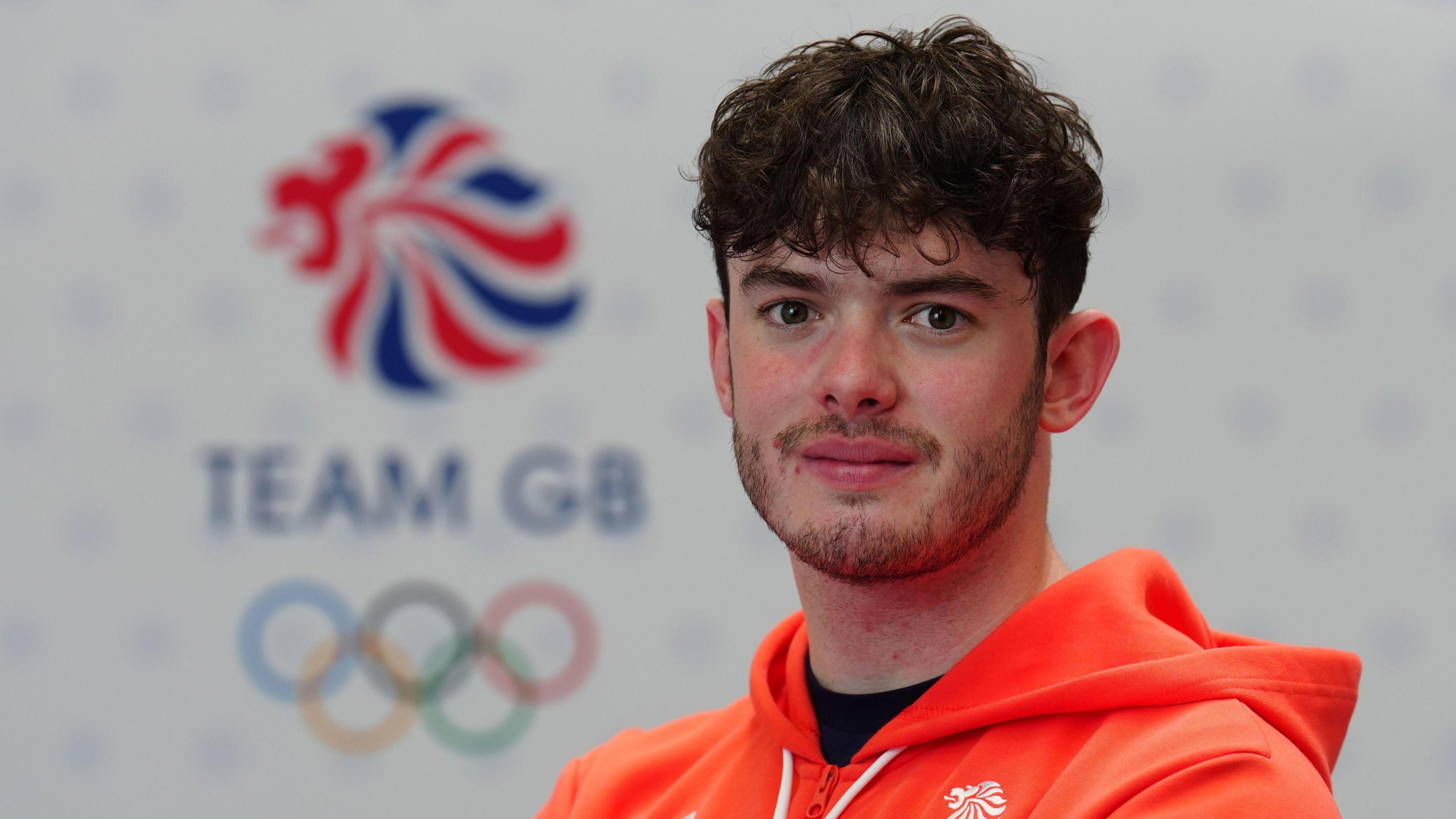 A head and shoulders view of Ed Lowe during a Team GB kitting out session for the Paris Olympics. He has dark brown hair with a fringe, a short beard and moustache and is wearing a red hoodie. In the background is a Team GB Olympic logo.