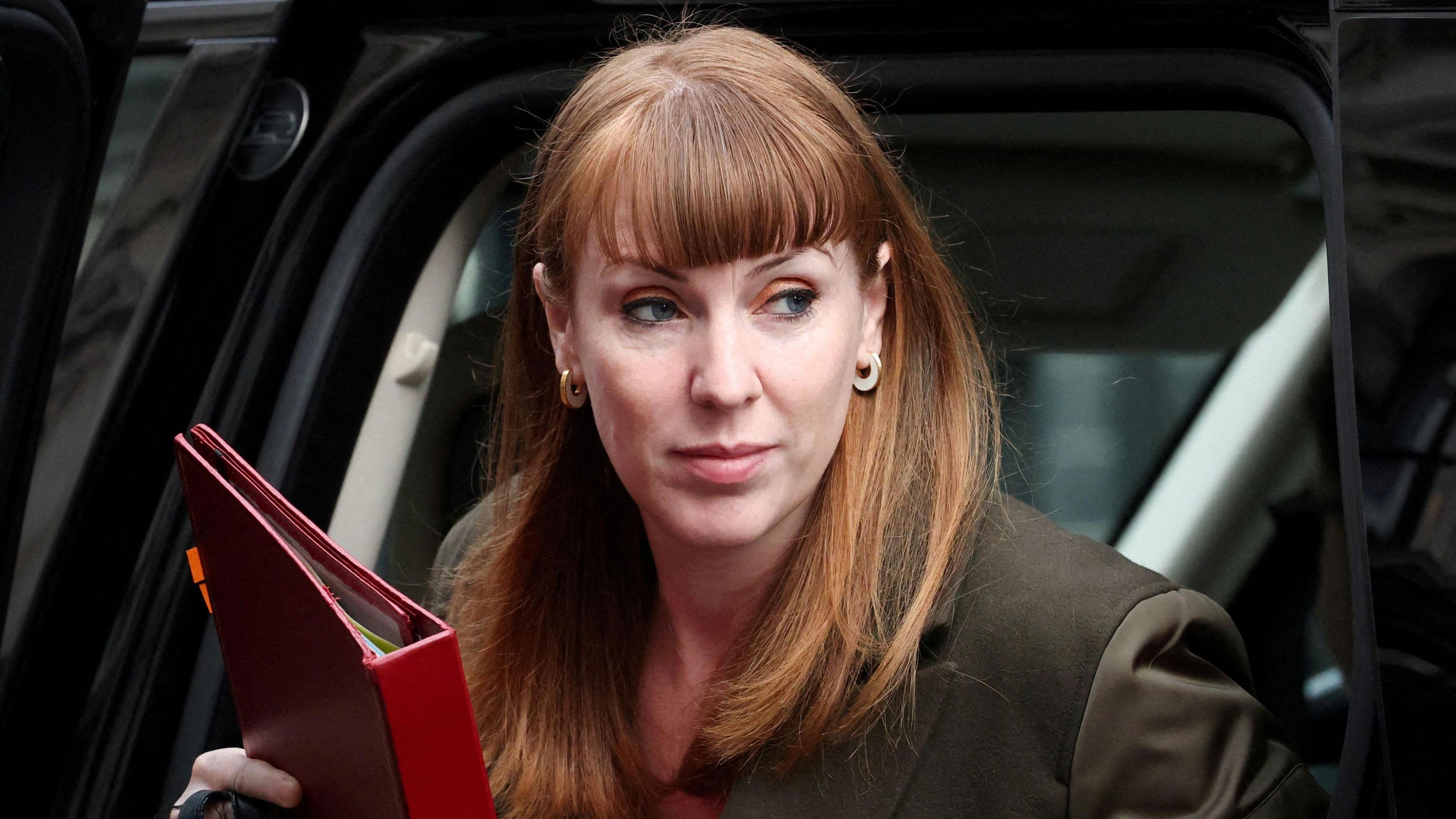 Angela Rayner - a woman with long red hair and wearing a suit - gets out of a black car and looks to her left while holding a red folder in her right hand.