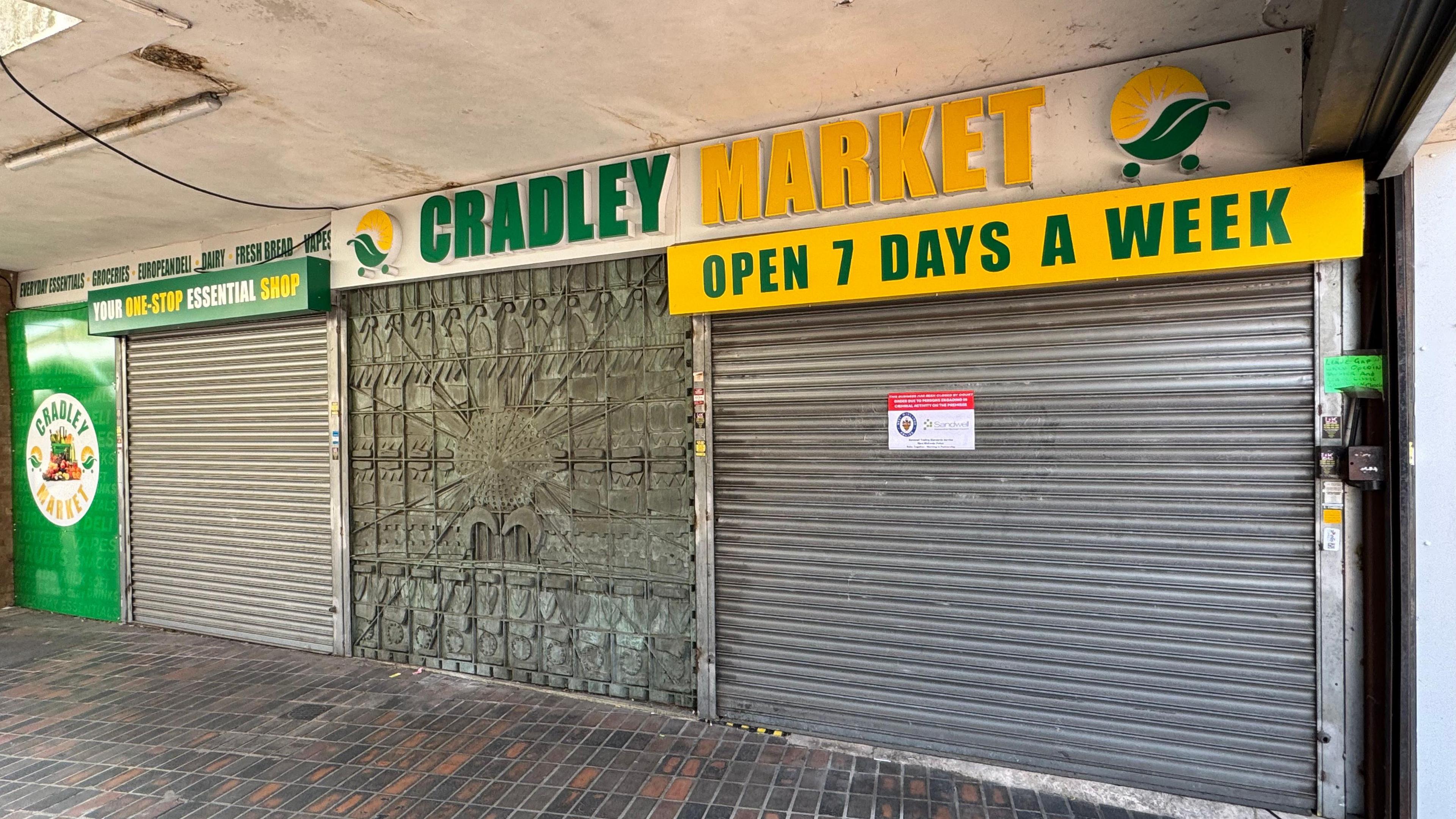 The frontage of the Cradley Market store, with green and yellow signage, with silver shutters that have been closed 