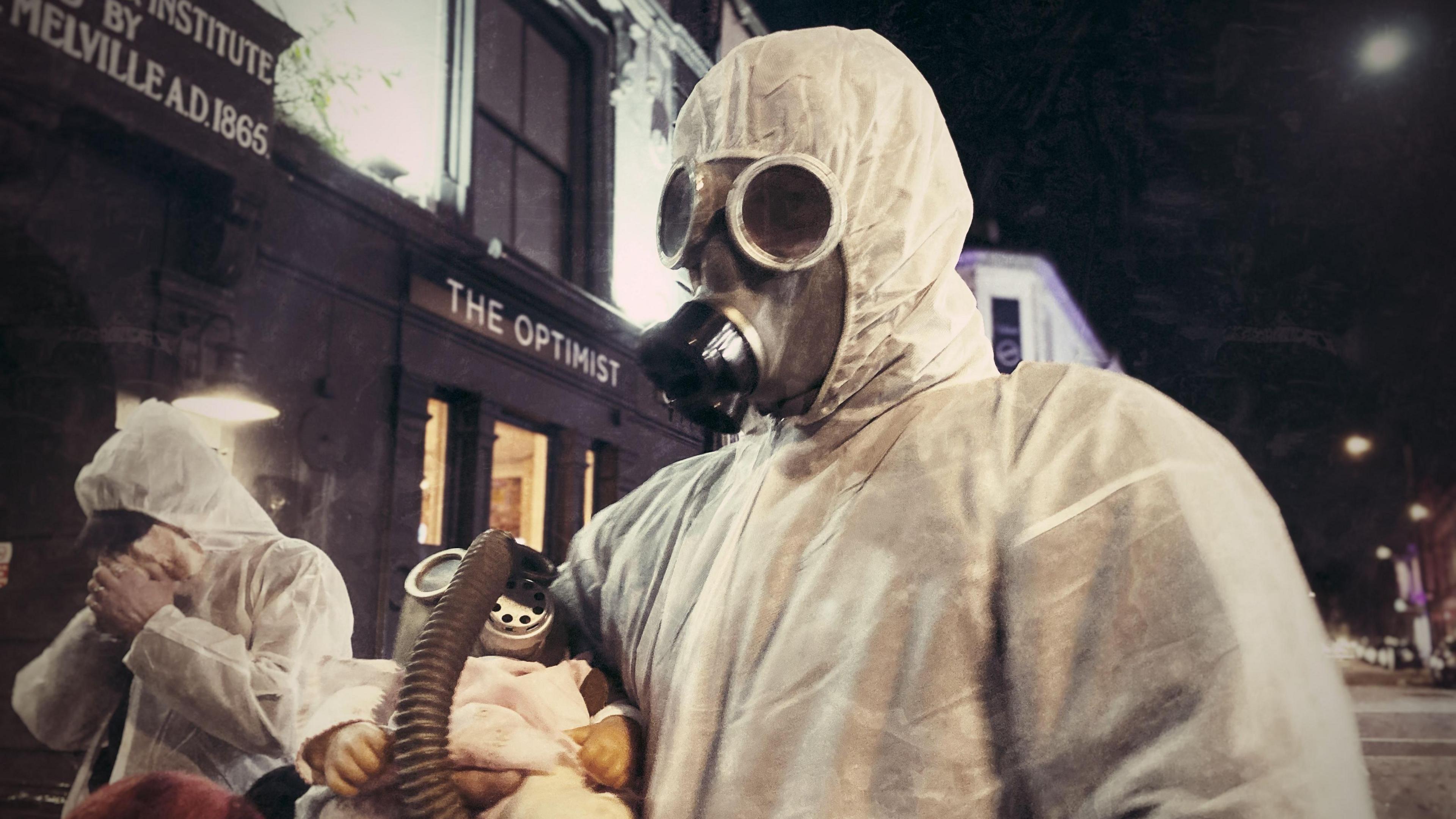 A person wearing a Hazmat suit and gas mask while holding a baby