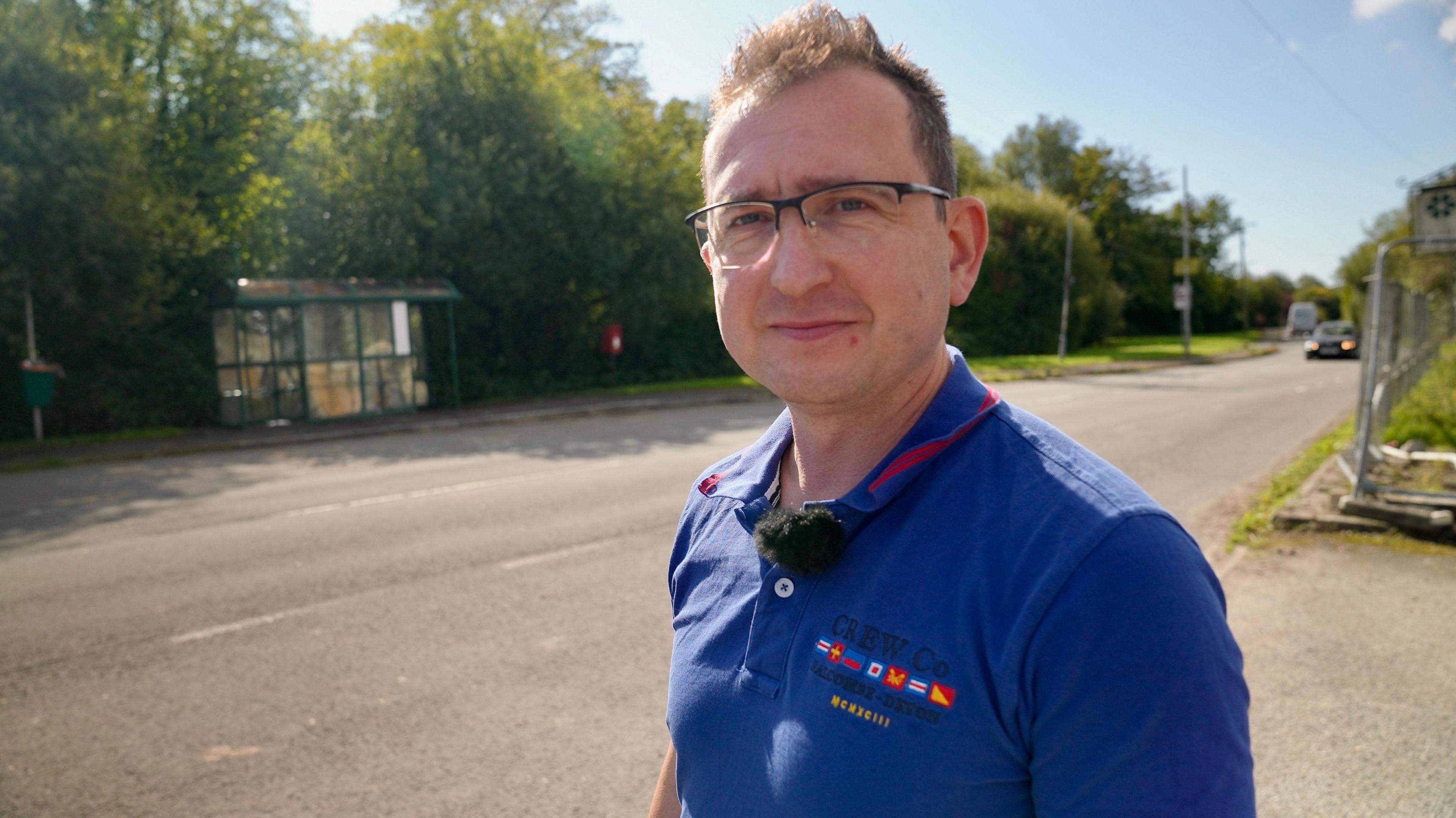 Lee Colvin is wearing glasses and a blue polo t-shirt staring at the camera, with the village of Peterstone behind him.
