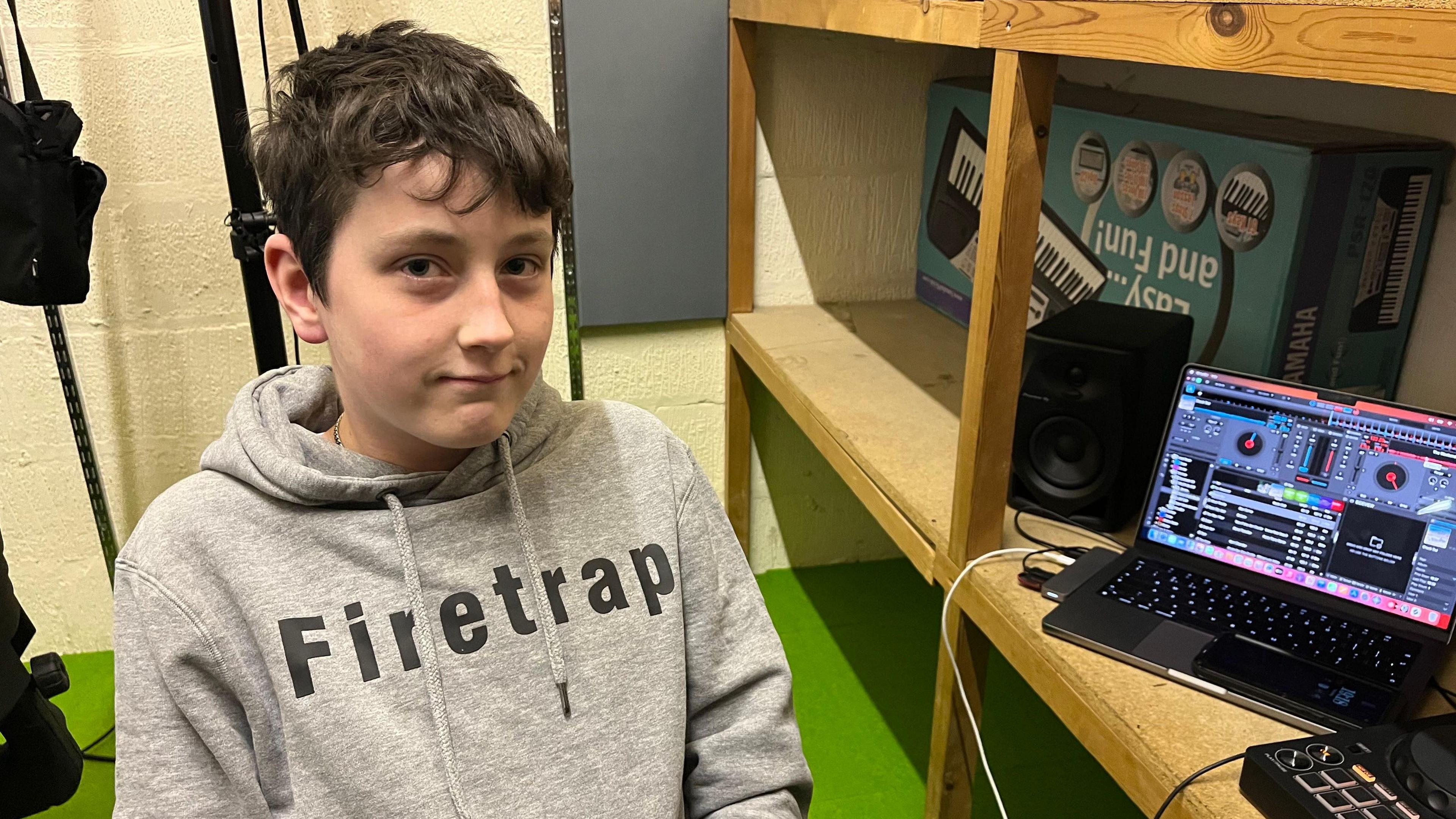 A young boy in a grey hoodie sits by a wooden desk with a laptop with DJ software on the computer.