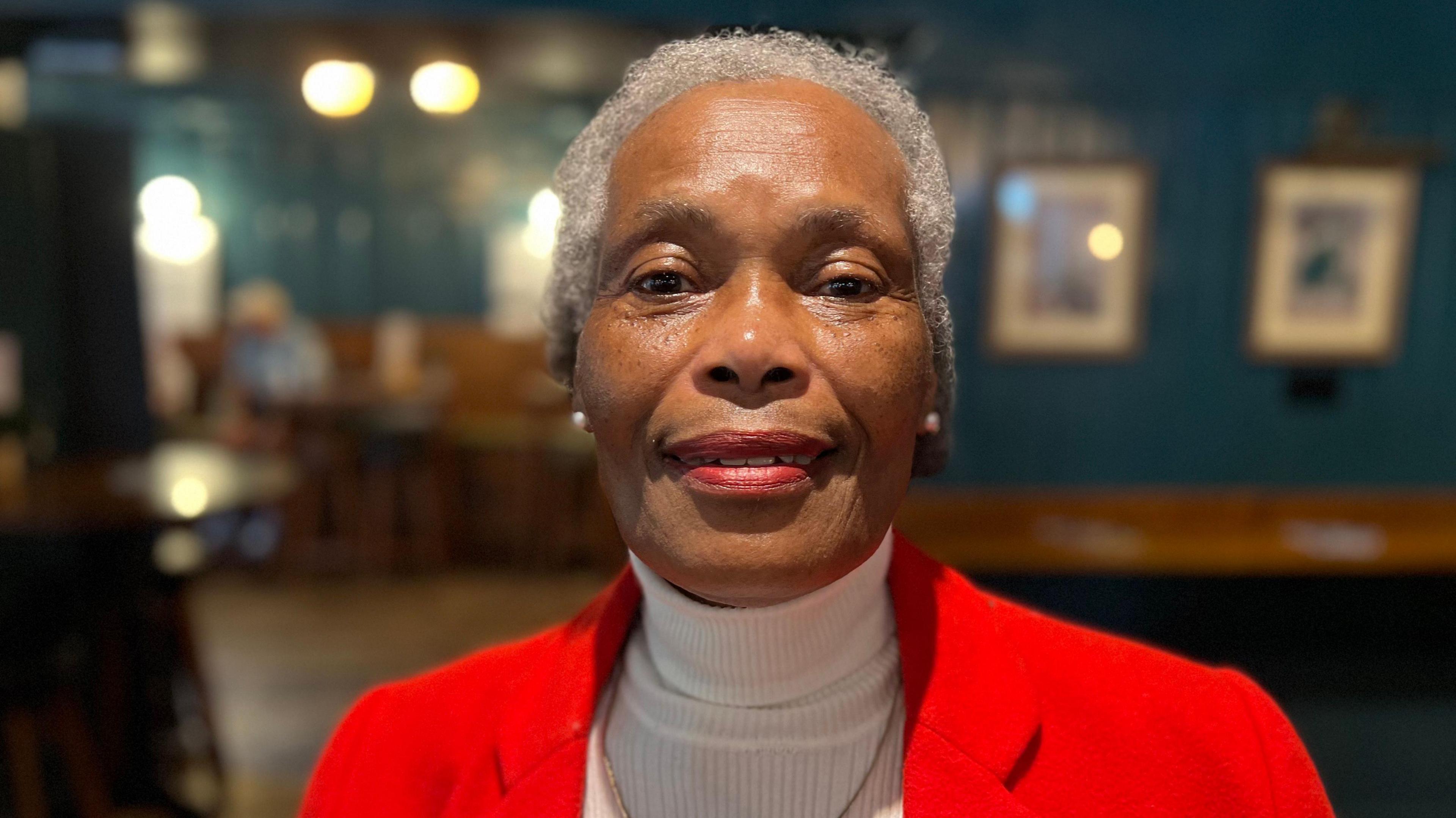 Joyce Morris-Wisdom is sitting in a pub and wearing a red blazer with a white turtle-neck underneath. She has short, grey hair and is smiling at the camera.