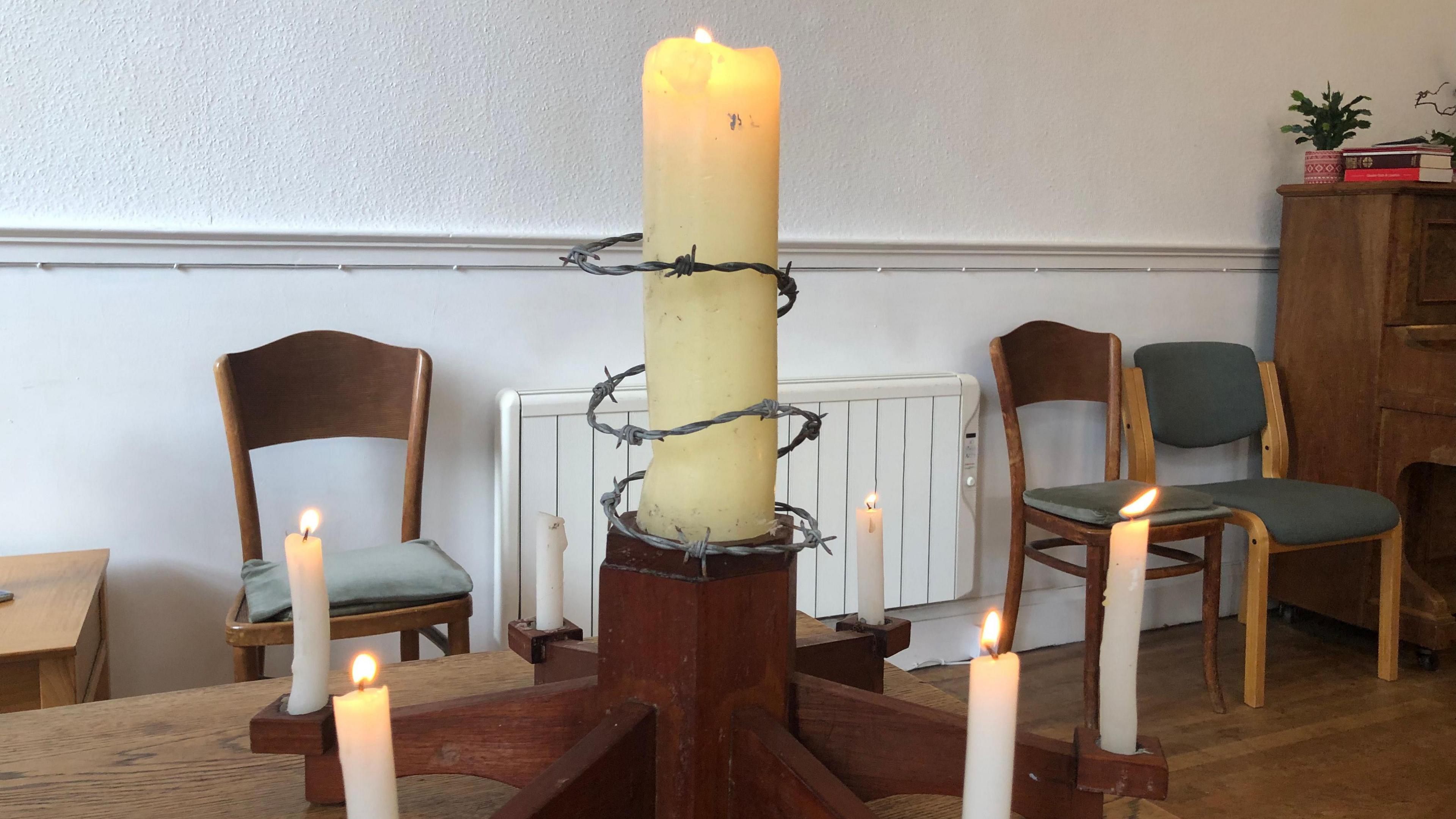 A large candle, with a thread of barbed wire around it, is in the centre of a wooden holder, with six smaller candles around the edge