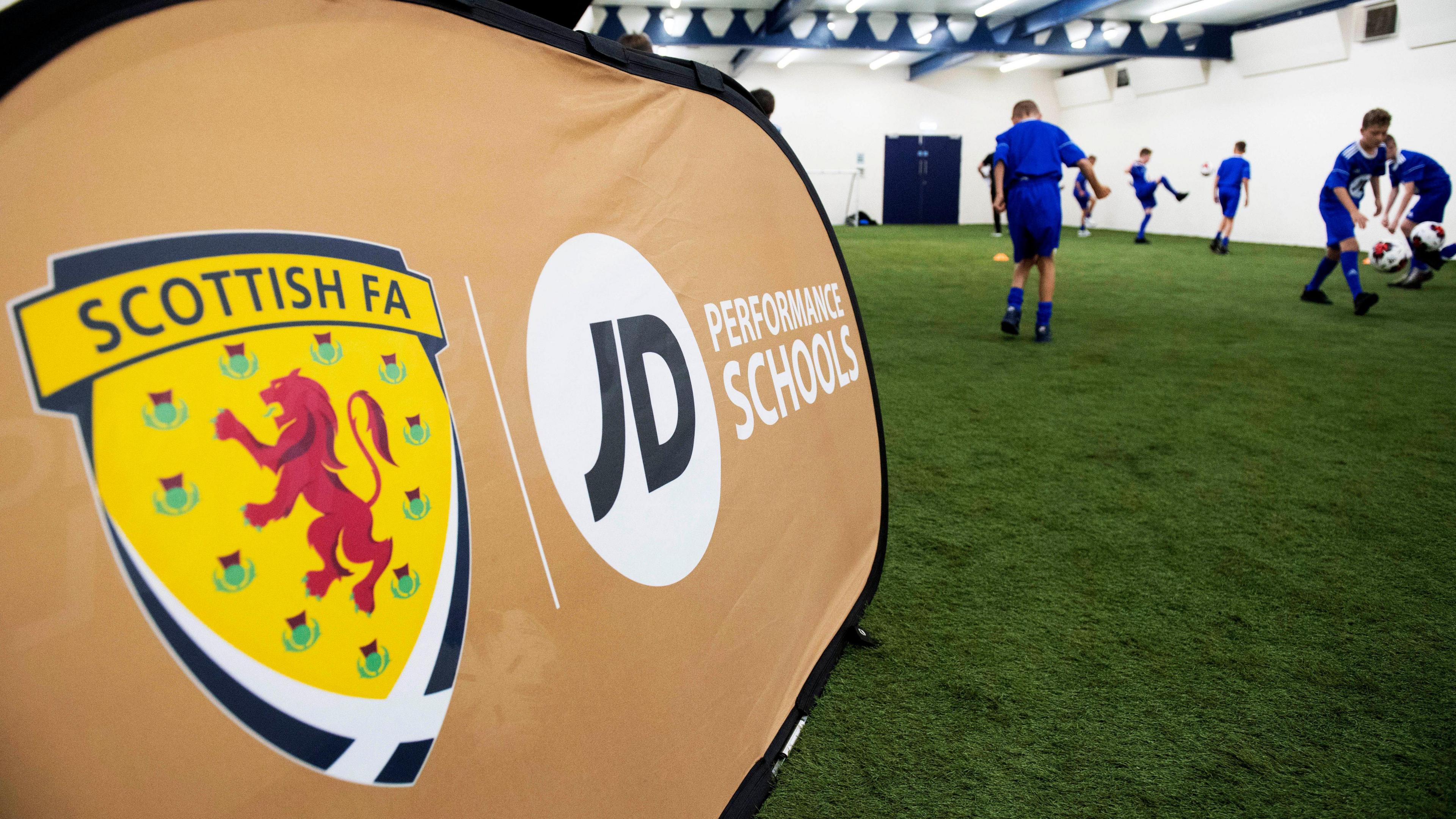 A banner advertising performance schools with the SFA logo. Boys in blue football strips are playing football alongside it in an indoor area.