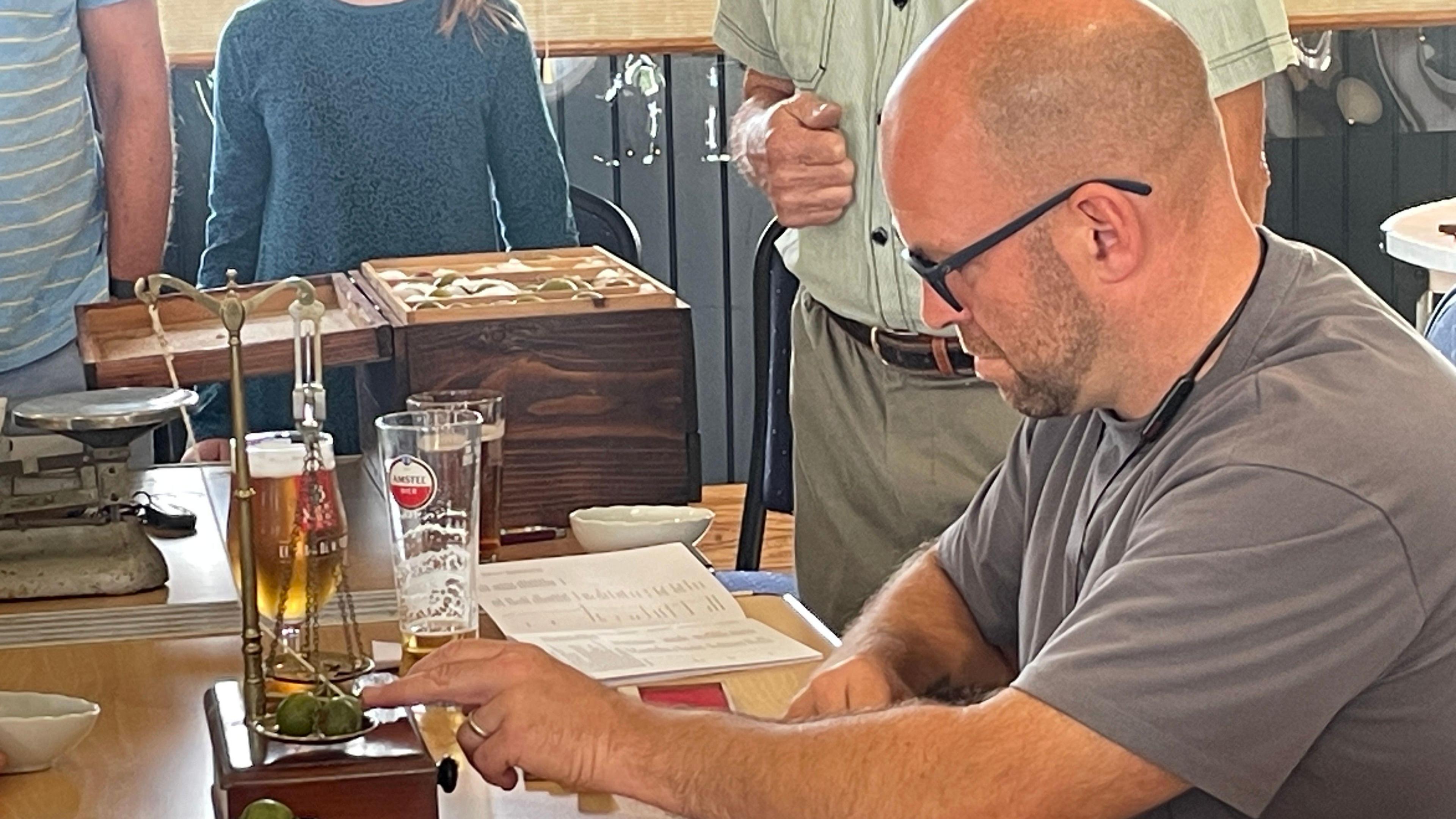A man wearing spectacles weighs a few gooseberries in traditional scales on a table. Some beer glasses, notebooks and boxes are in the background with people standing around him.
