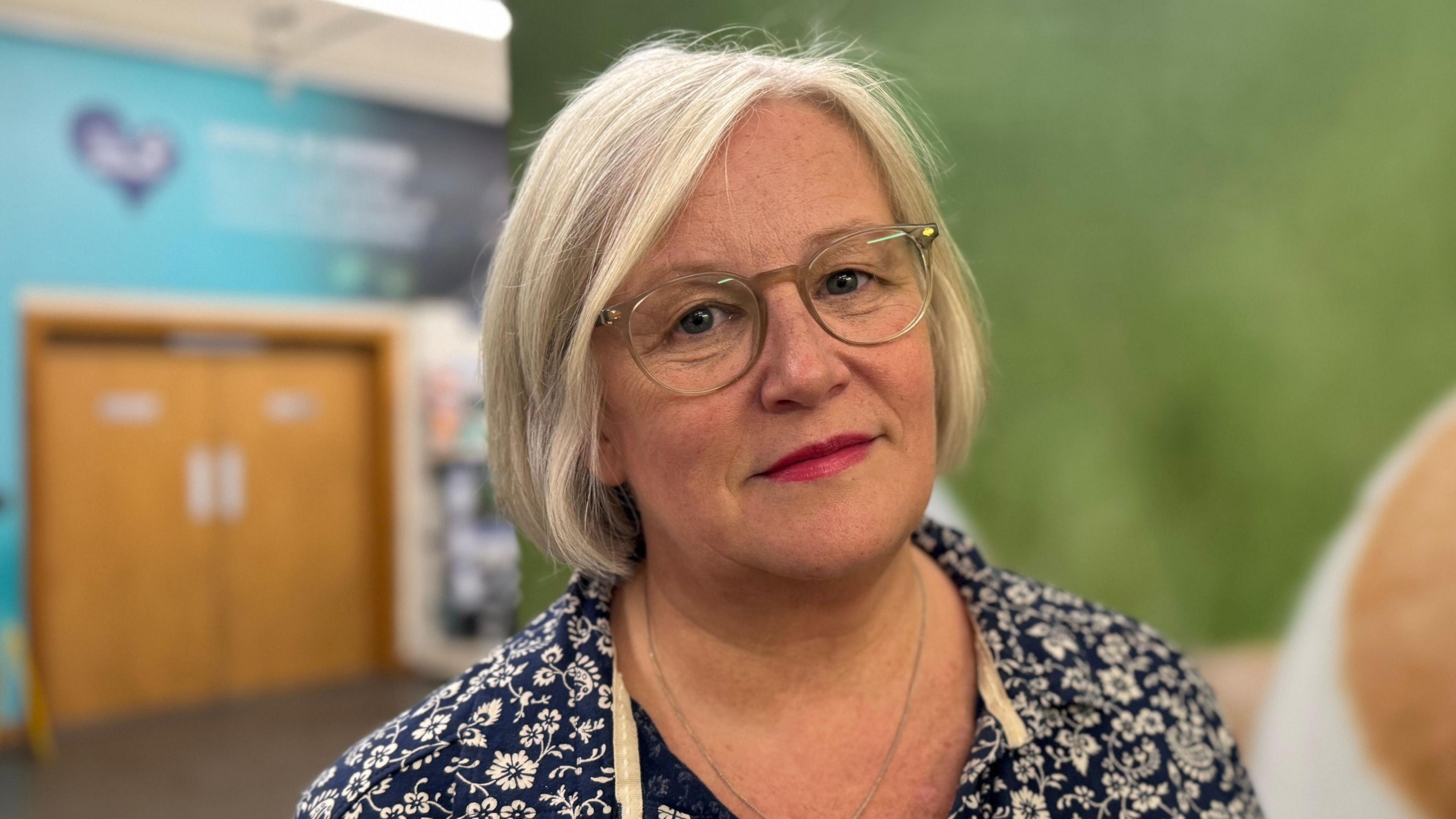 Heather Weir stands in the middle of the USPCA shop in Newry in front of a large wall display. She has short, blonde hair and is wearing a pair of green plastic glasses. She is wearing a blue and white flower shirt and is looking into the camera with a neutral expression on her face.