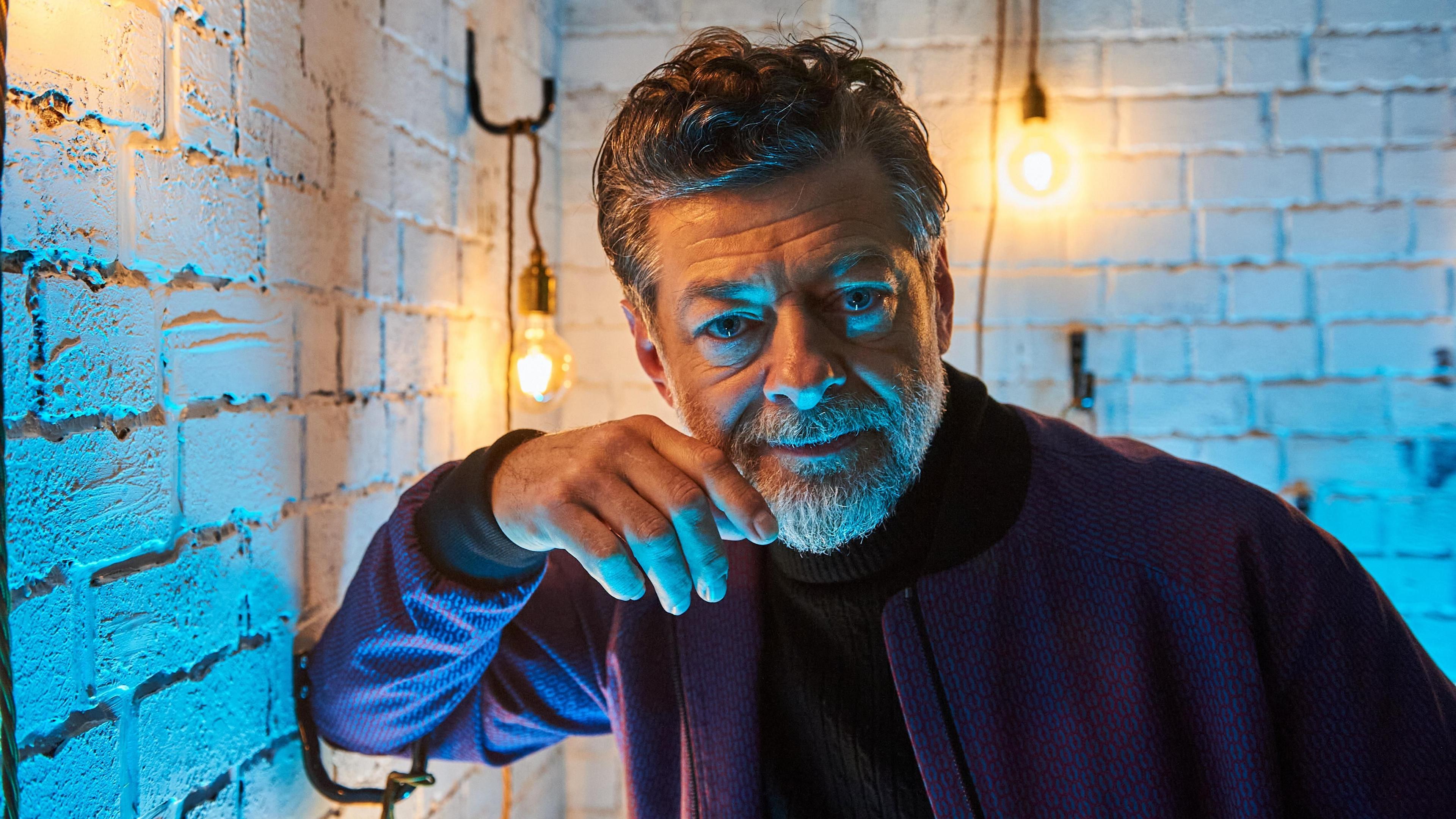 Andy Serkis is stood in a dimly lit room. Blue light is illuminating the features on his face. Behind him he is back-lit by golden light from exposed light bulbs. 