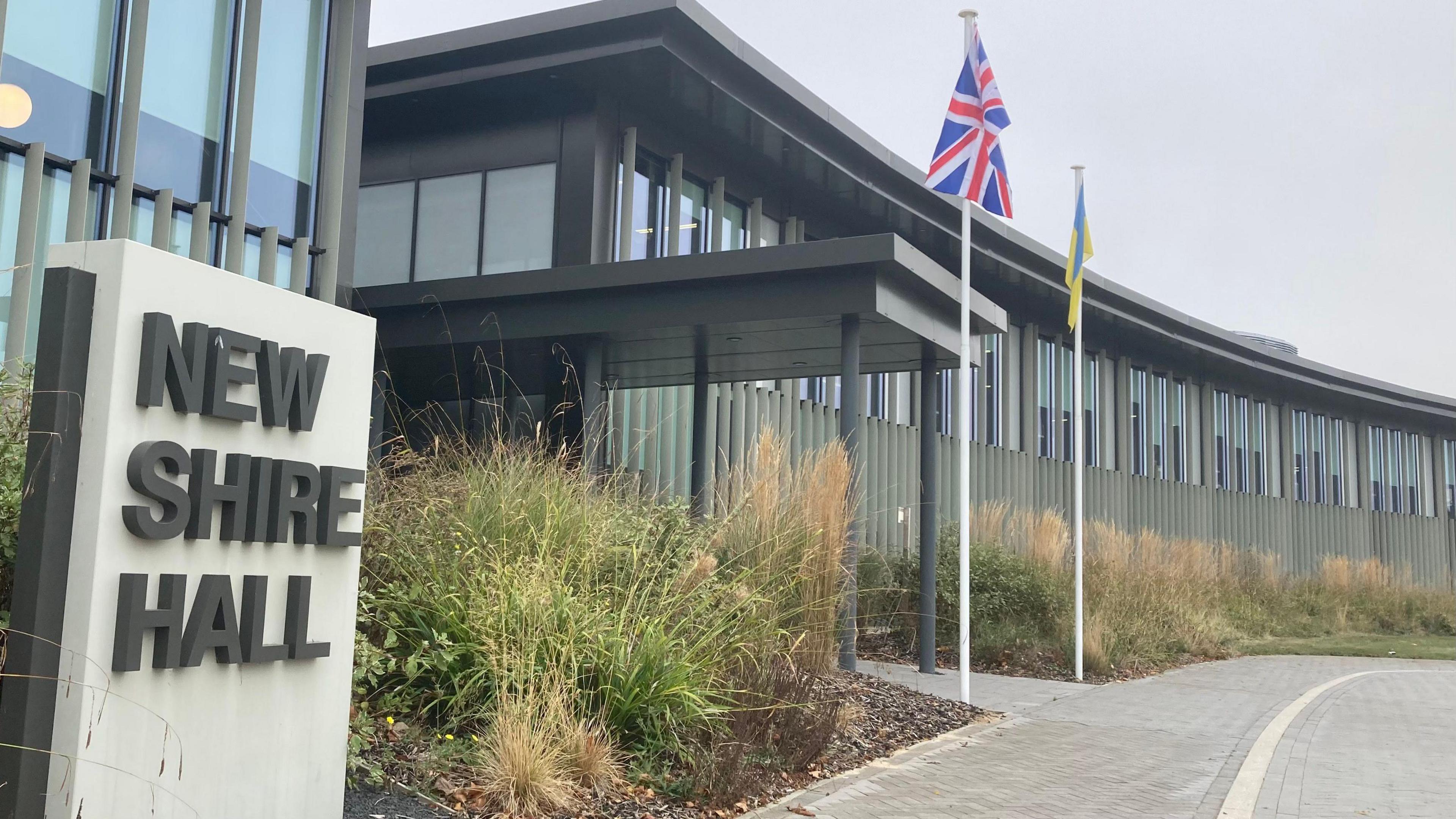 The entrance to Cambridgeshire County Council's New Shire Hall headquarters. It is a long, sleek, grey building with tall windows and it looks modern. Grass has been allowed to grow tall in length out the front, which also has Union Jack and Ukraine flags on tall poles. There is a sign that says "New Shire Hall".