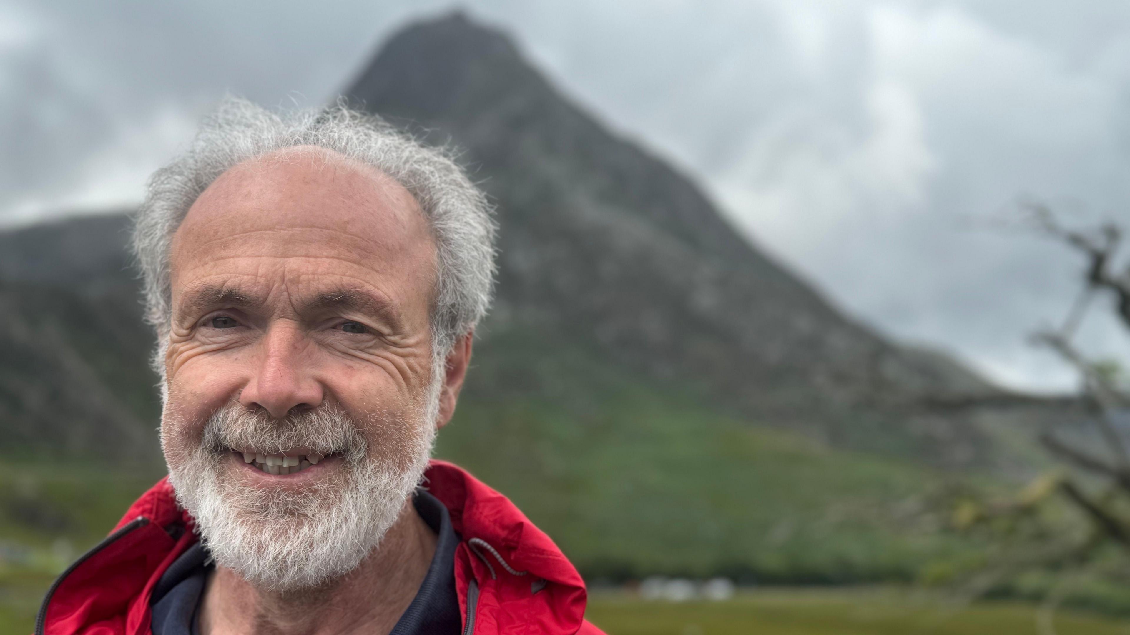 Portrait of Chris Lloyd with Tryfan in the background. He has a grey beard and grey hair and is wearing a red jacket and navy top