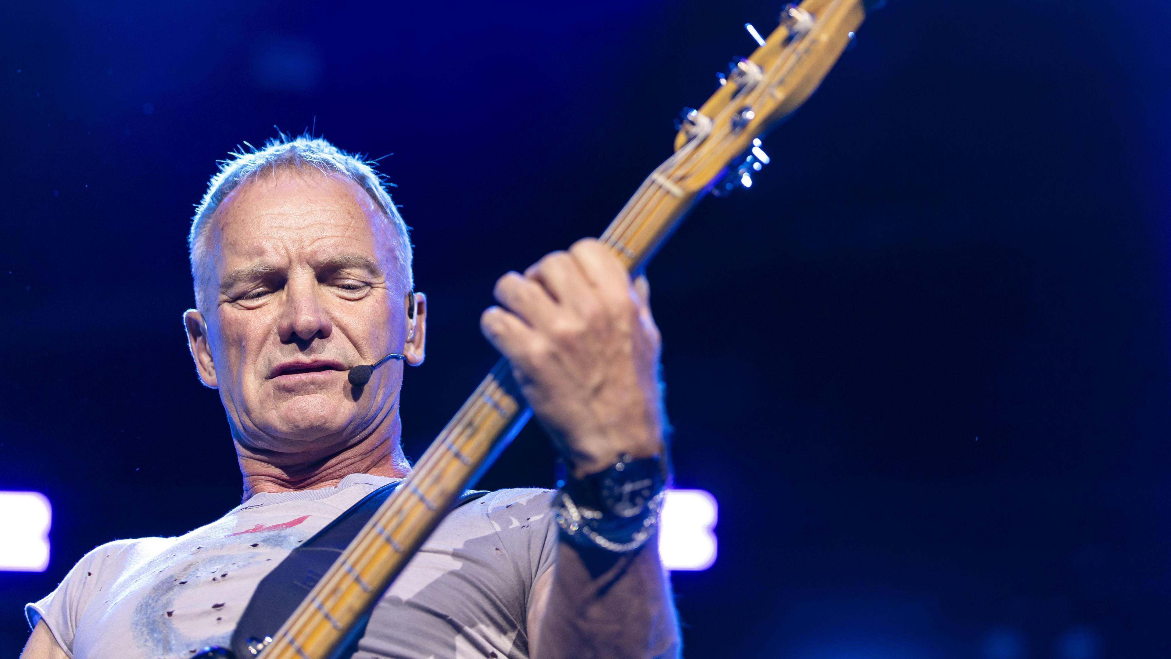 A man holds a guitar on stage, wearing a grey-t-shirt. He has short, grey/white hair and is looking down at the guitar.