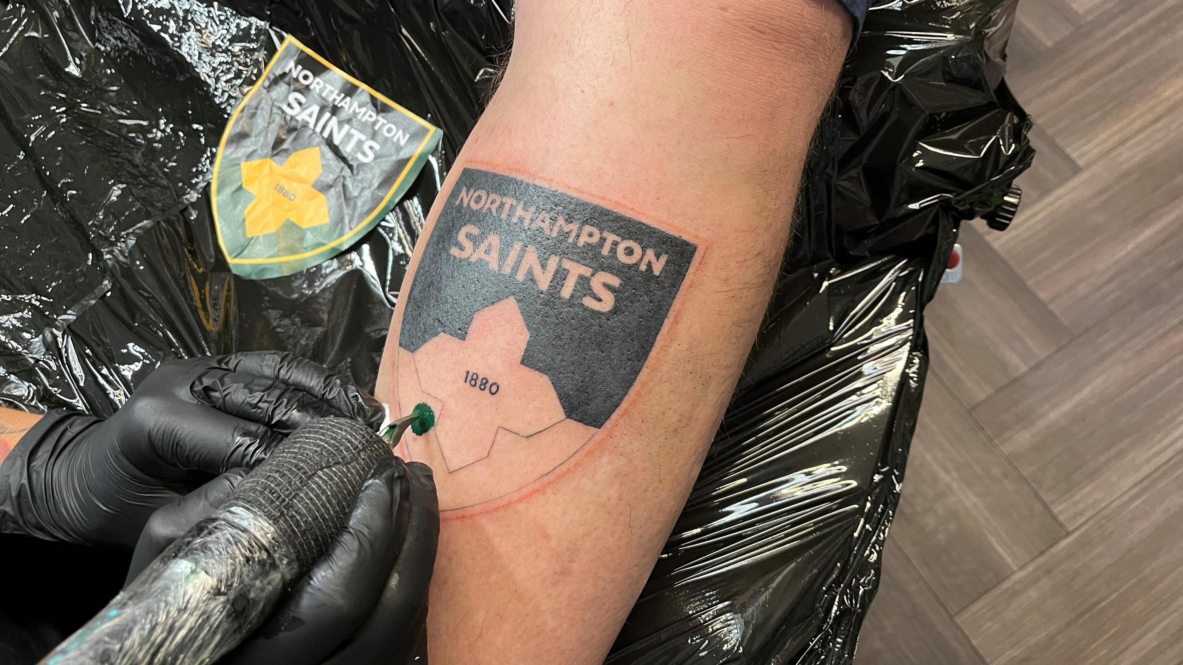 A close-up of Simon Stacey's arm with a showing half-finished Northampton Saints tattoo and the gloved hands of a tattoo artist holding a needle