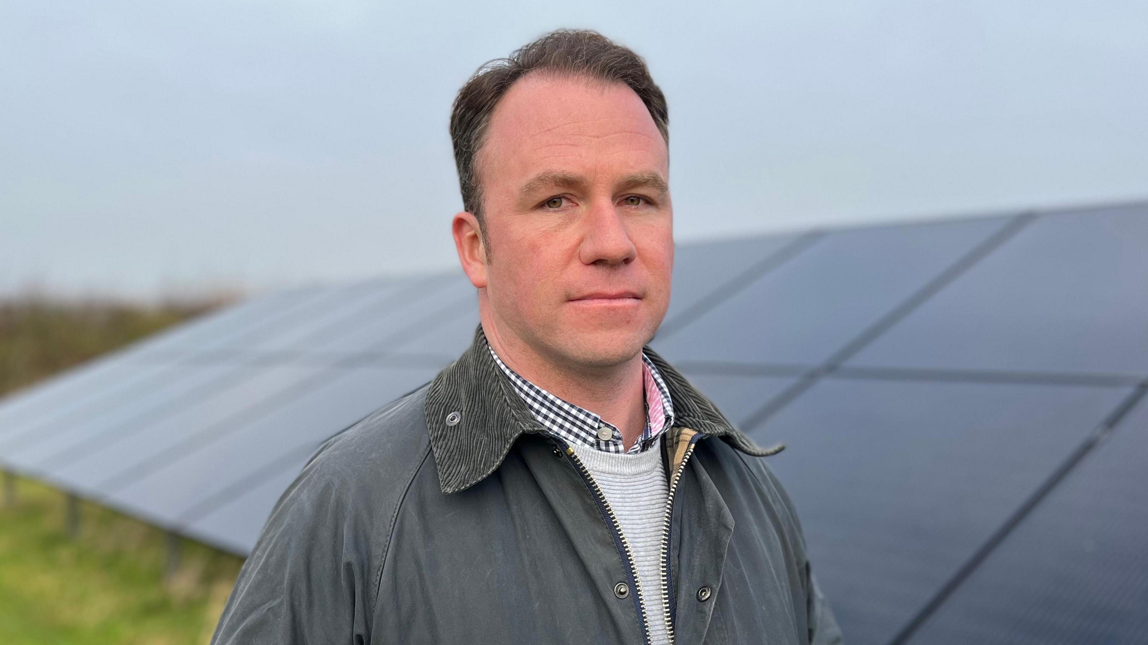 Richard Gill wearing a grey jacket, stands in front of some solar panels.