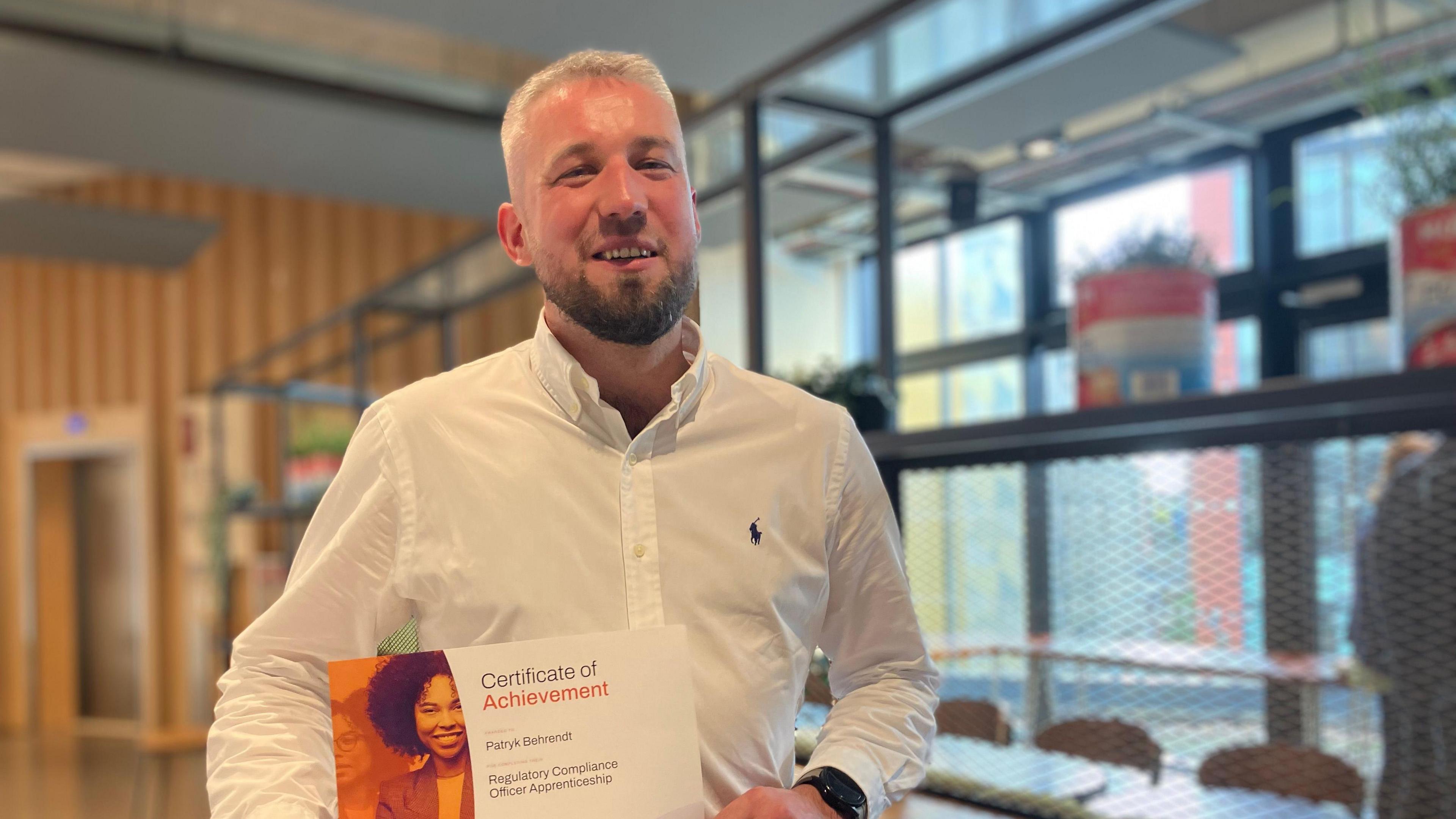 Patryk Behrendt holding up his certificate, wearing a plain white shirt in a conference room.